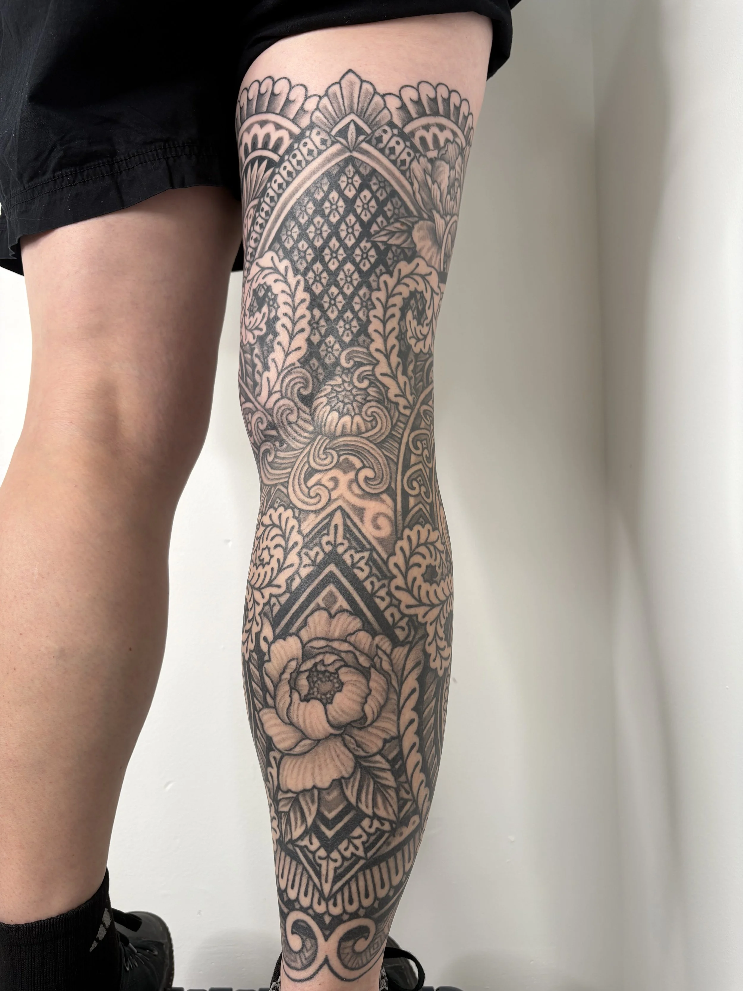 A person's leg is showcased with a detailed black and gray tattoo sleeve featuring floral patterns, geometric designs, and ornate elements.