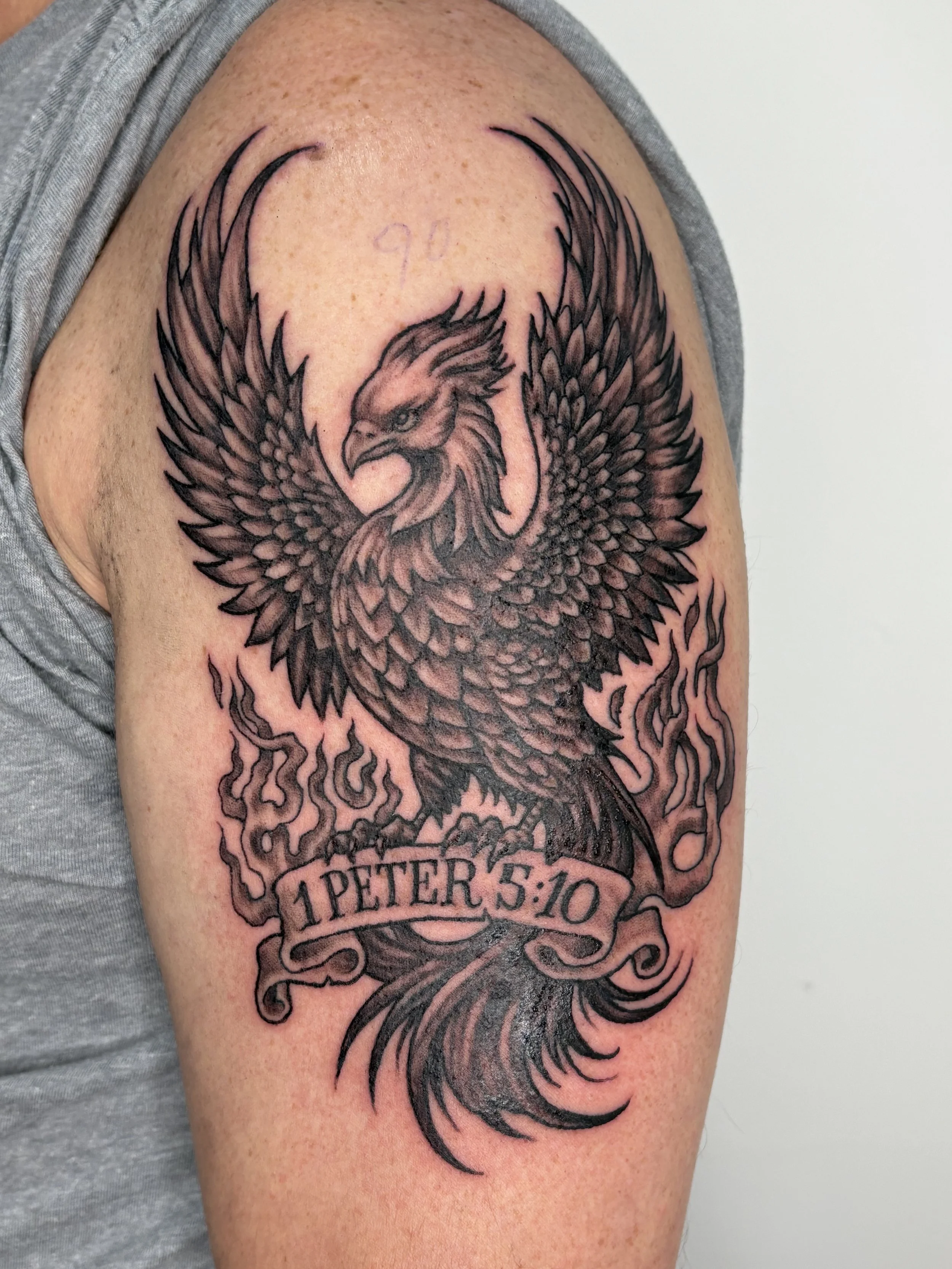Black and gray tattoo of a phoenix with outstretched wings on a person's upper arm, below the lion with a banner reading "1 PETER 5:10". State Of The Art Tattooing Winchester Virginia  