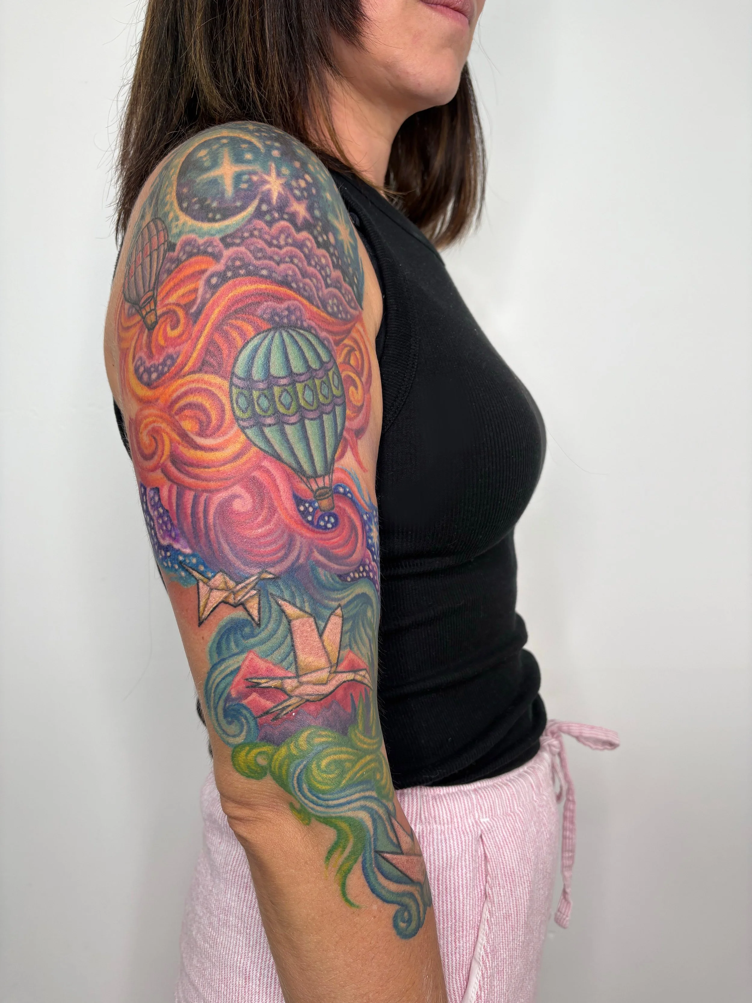 Close-up of a woman's upper arm, showing a colorful tattoo of hot air balloons, clouds, stars, and swirling patterns. State Of The Art Tattooing Winchester Virginia 