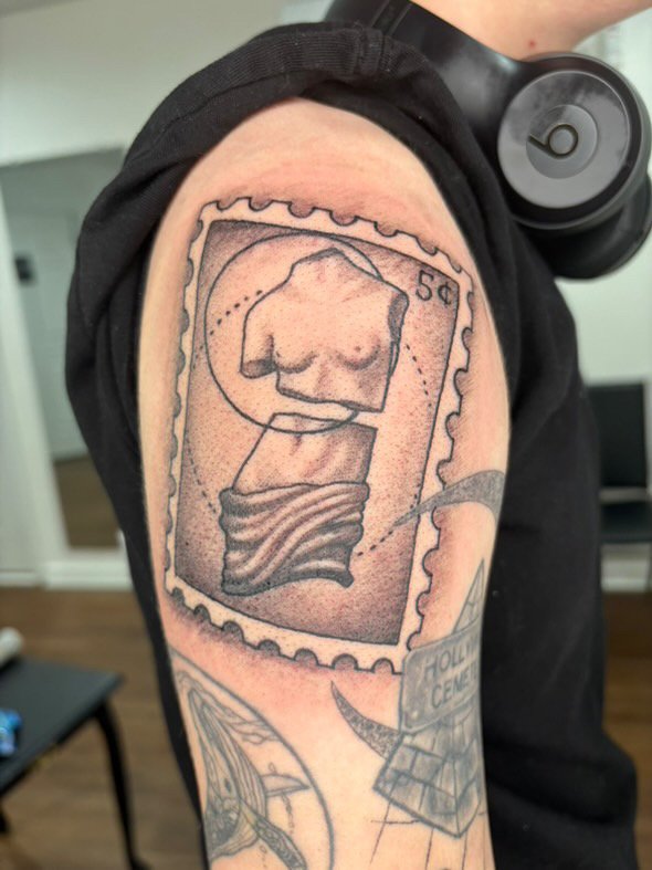Tattoo of a vintage camera with a woman's chest on the film area and a partially visible sidewalk sign reading 'HOLI CENTA' on the person's upper arm.