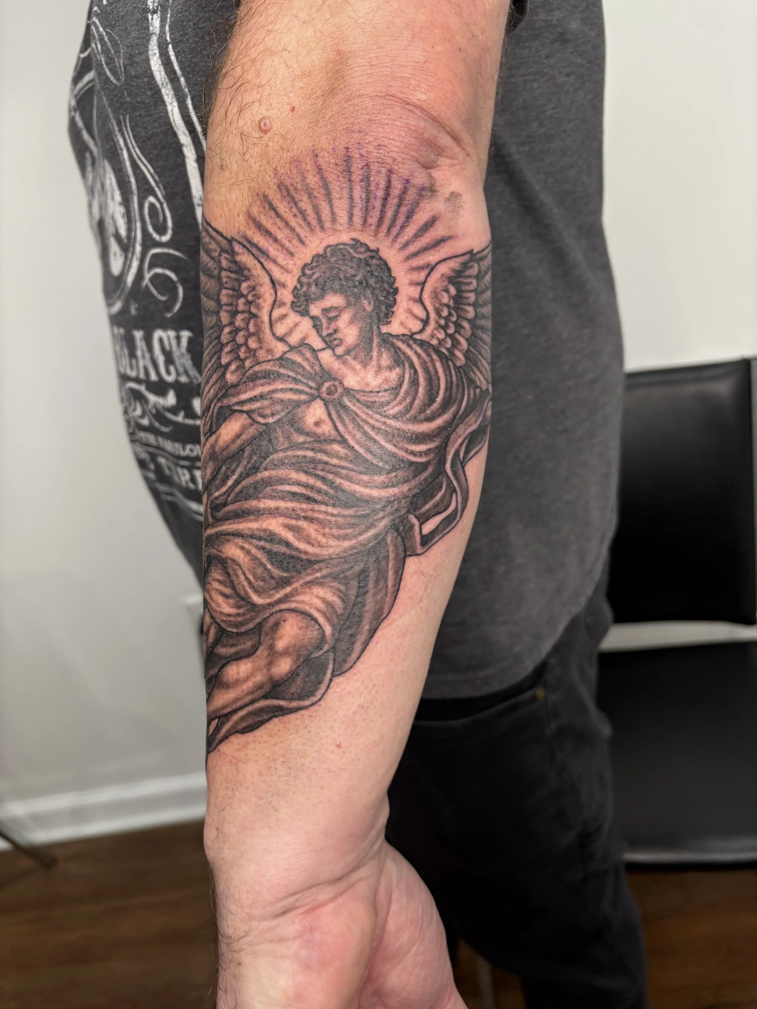 Close-up of a person's arm with a black ink tattoo of an angel with curly hair, wings, and a flowing robe on the upper arm. State Of The Art Tattooing Winchester Virginia 