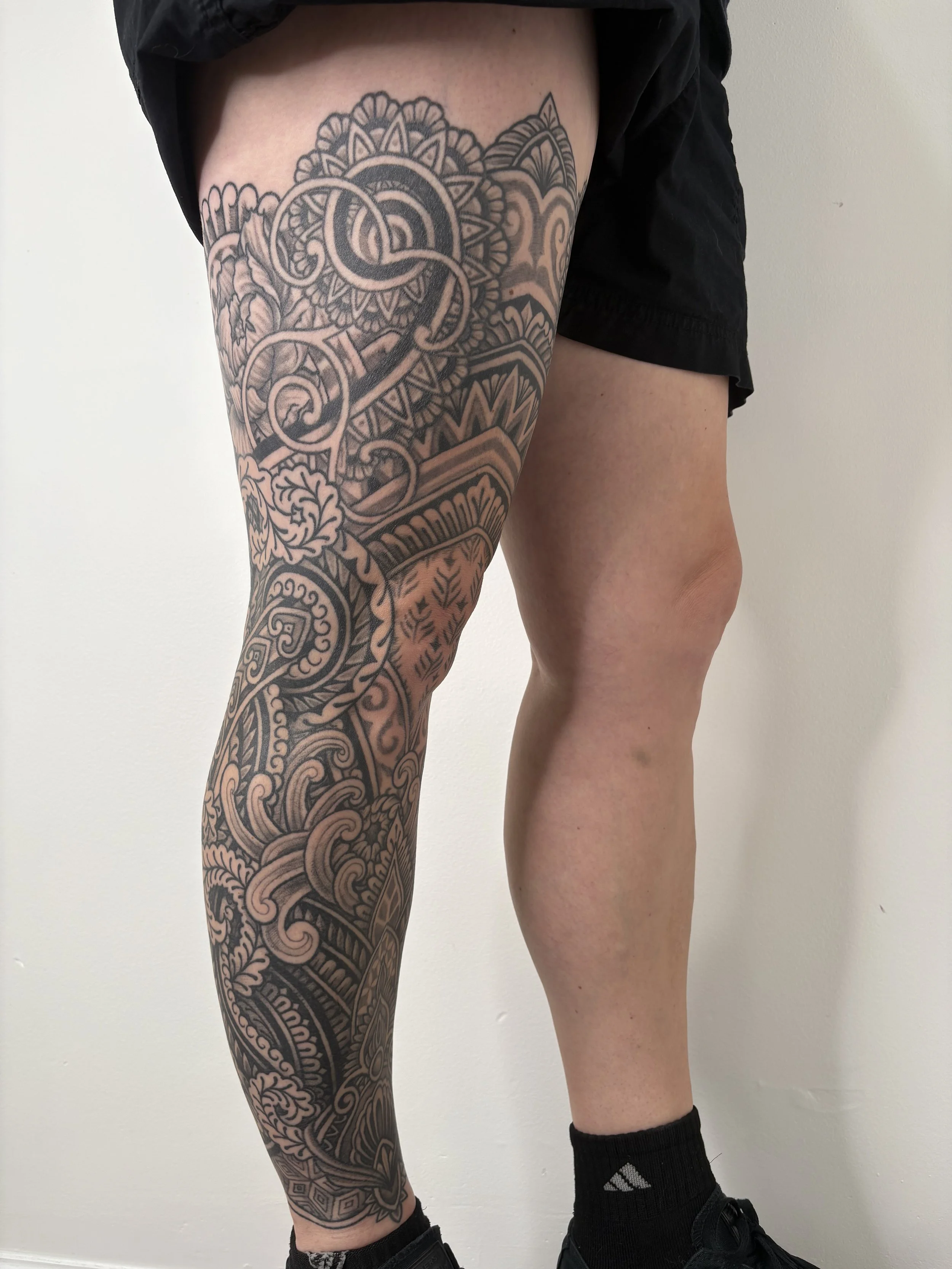 A person's leg with an intricate black tattoo extending from the thigh to the ankle, with a second person's leg visible beside it, wearing black socks and sneakers, against a plain white wall.