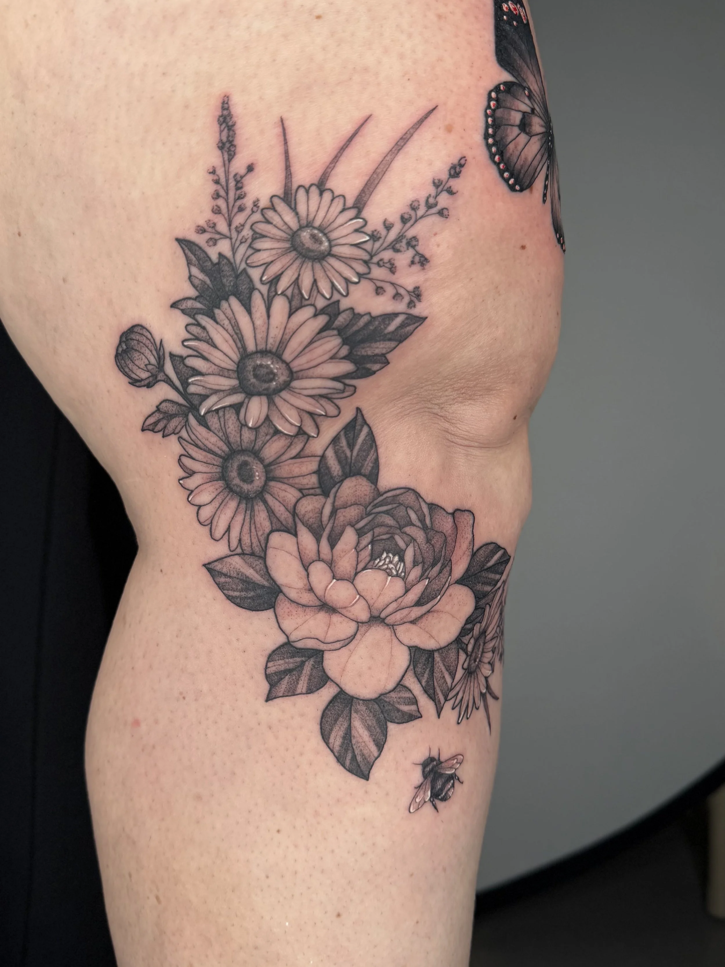 Close-up of a person's shoulder and upper arm with a black and gray floral tattoo featuring daisies, roses, leaves, and butterflies. State Of The Art Tattooing Winchester Virginia 