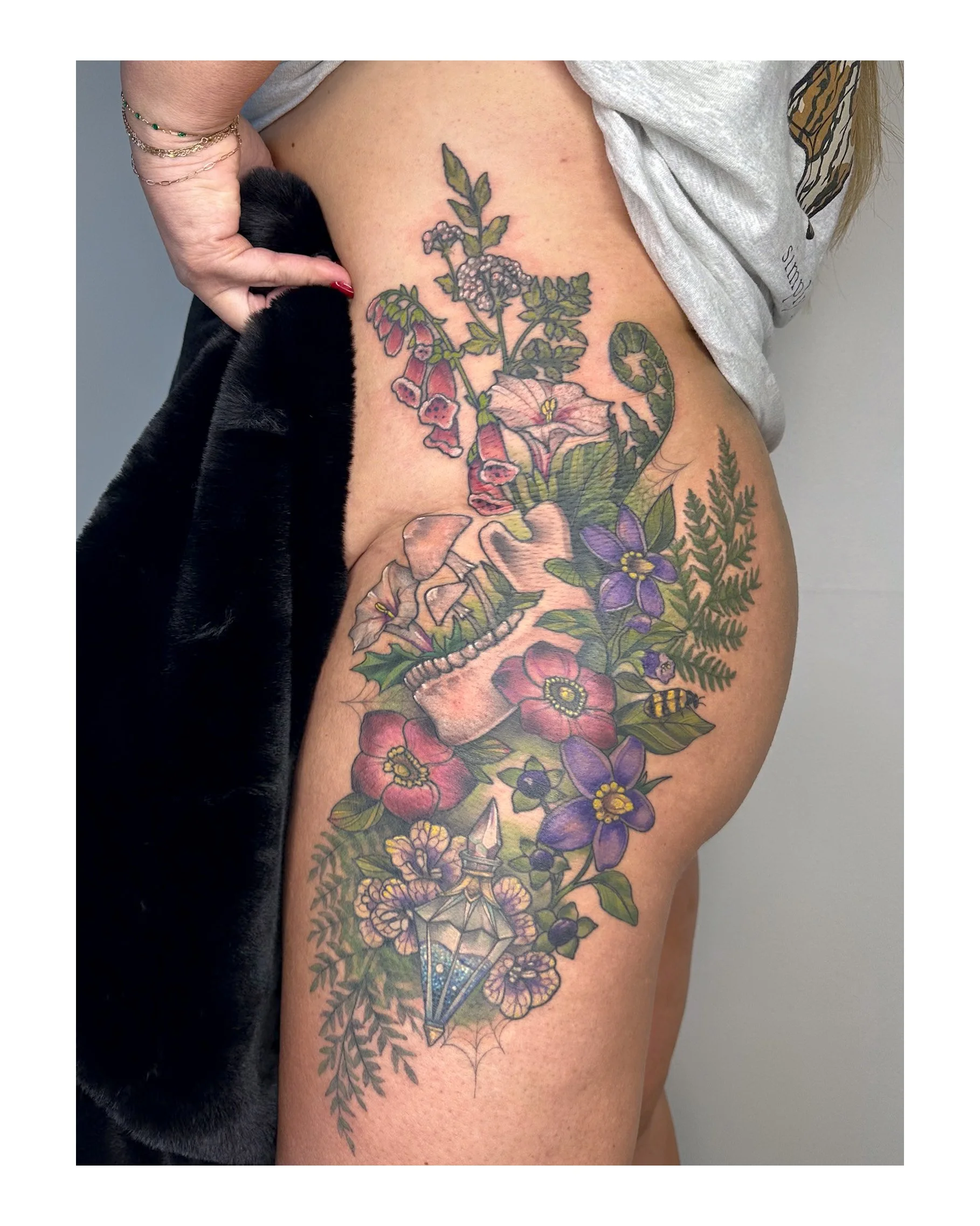 A person's upper thigh with a colorful floral tattoos, featuring various flowers, leaves, and a skull, partially covered by a gray T-shirt.
