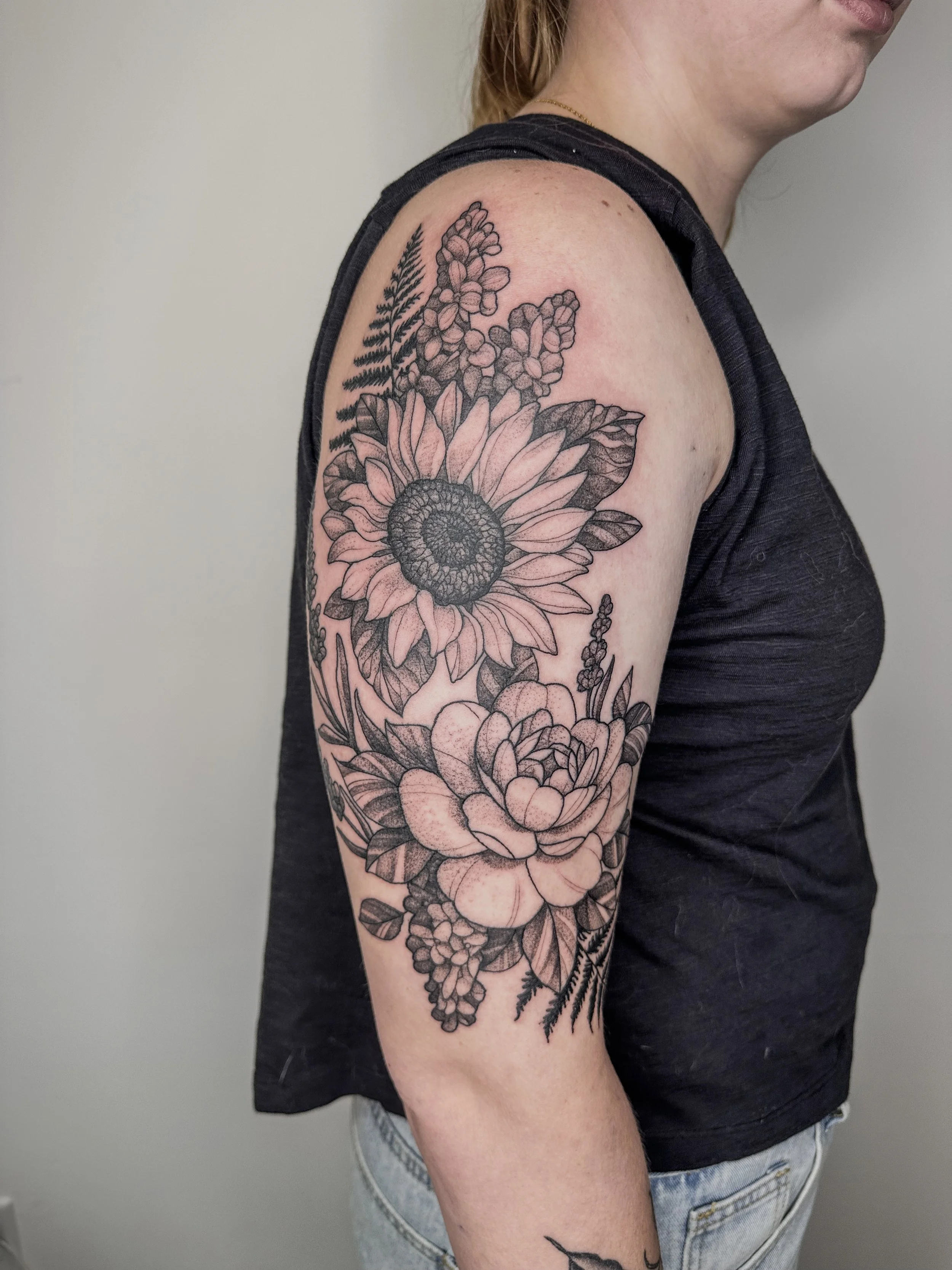 A woman with a large floral black ink tattoo on her upper arm and shoulder, featuring detailed sunflowers, ferns, and other botanical elements.