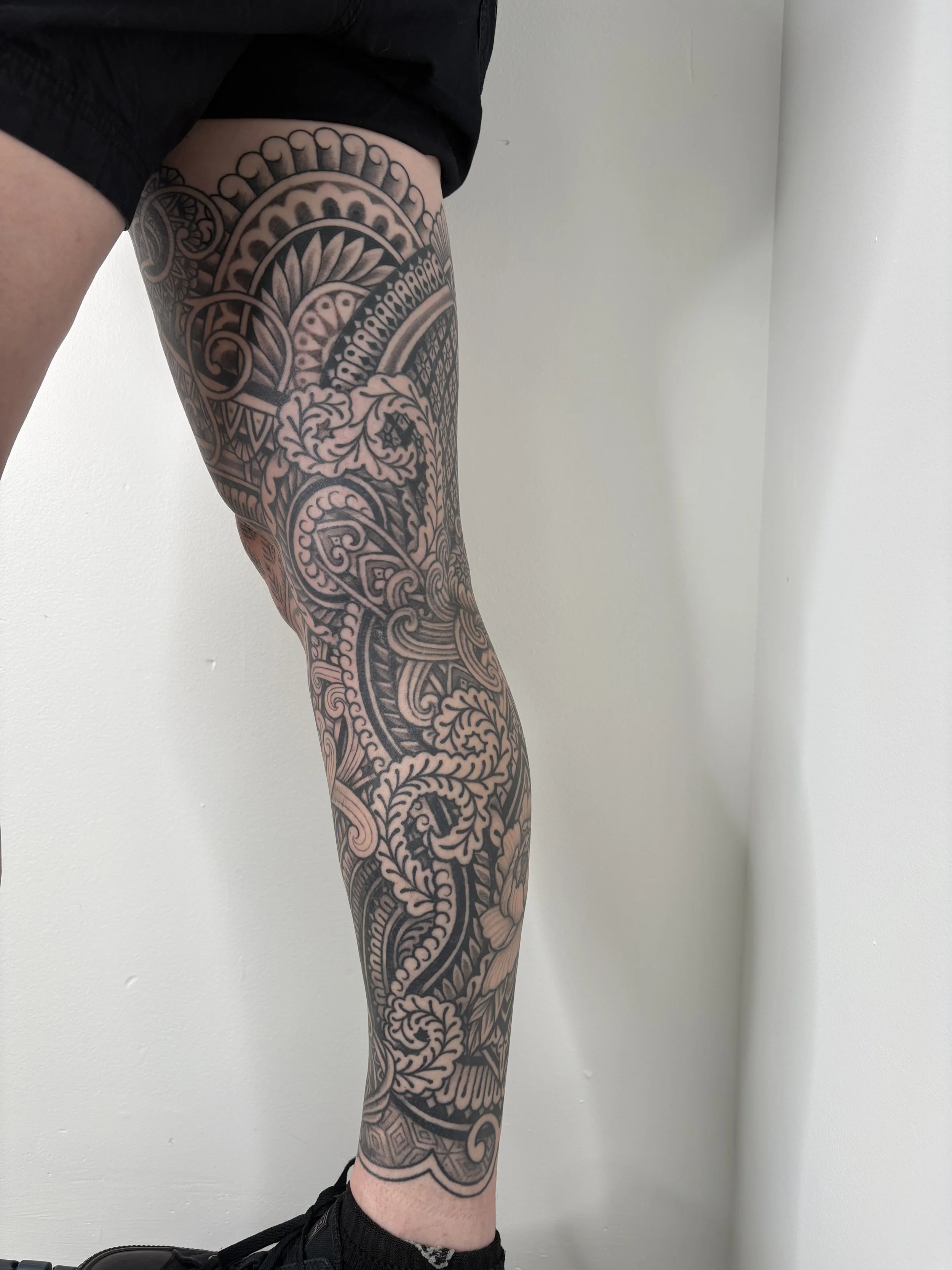 A person with a large, custom ornamental intricate black tattoo covering their leg, standing against a plain white wall, wearing black shorts and black sneakers. State Of The Art Tattooing Winchester Virginia 
