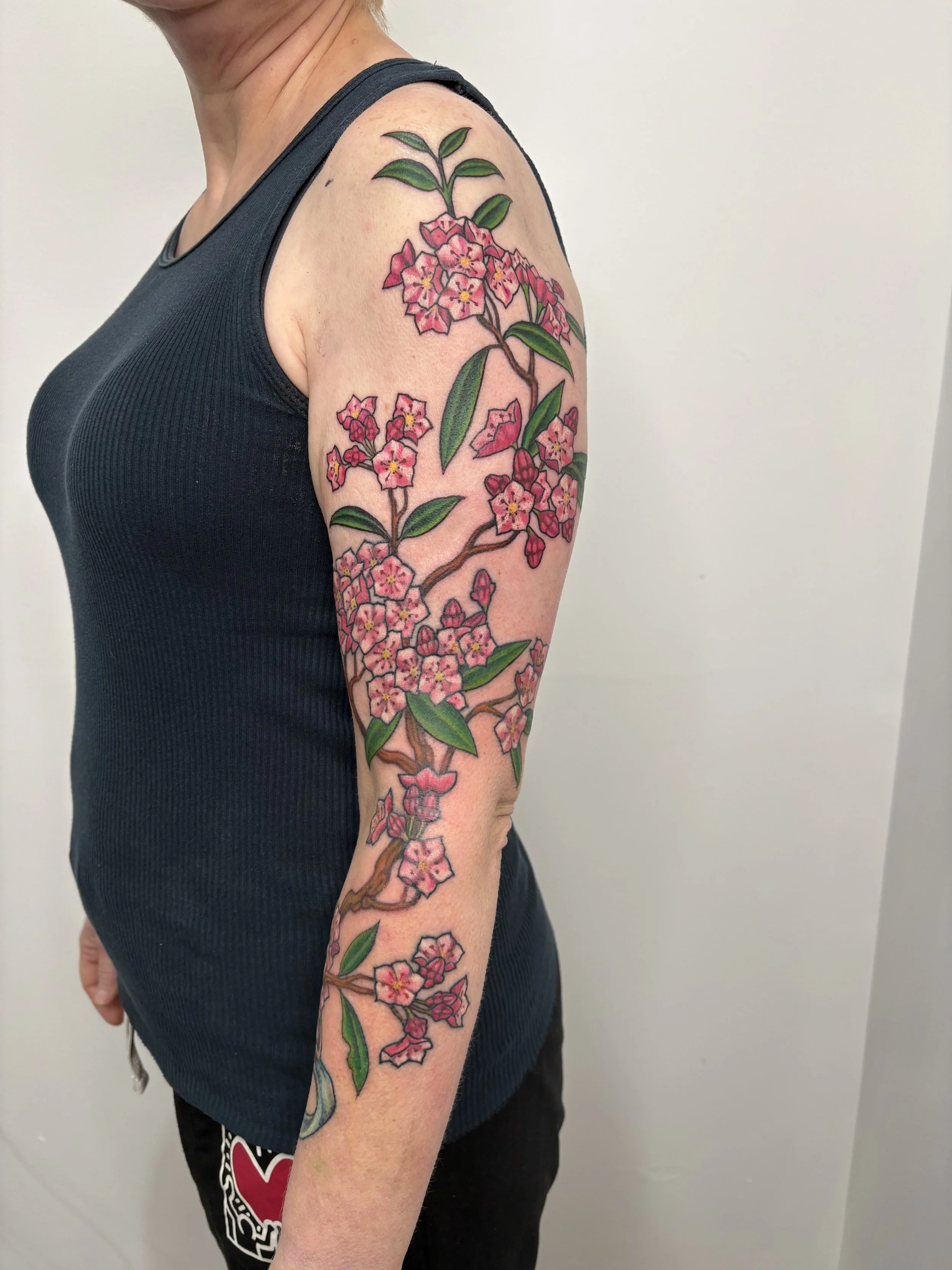 A person's upper arm and shoulder with a colorful floral tattoo featuring pink flowers with yellow centers and green leaves, extending from the shoulder down the arm. State Of The Art Tattooing Winchester Virginia 