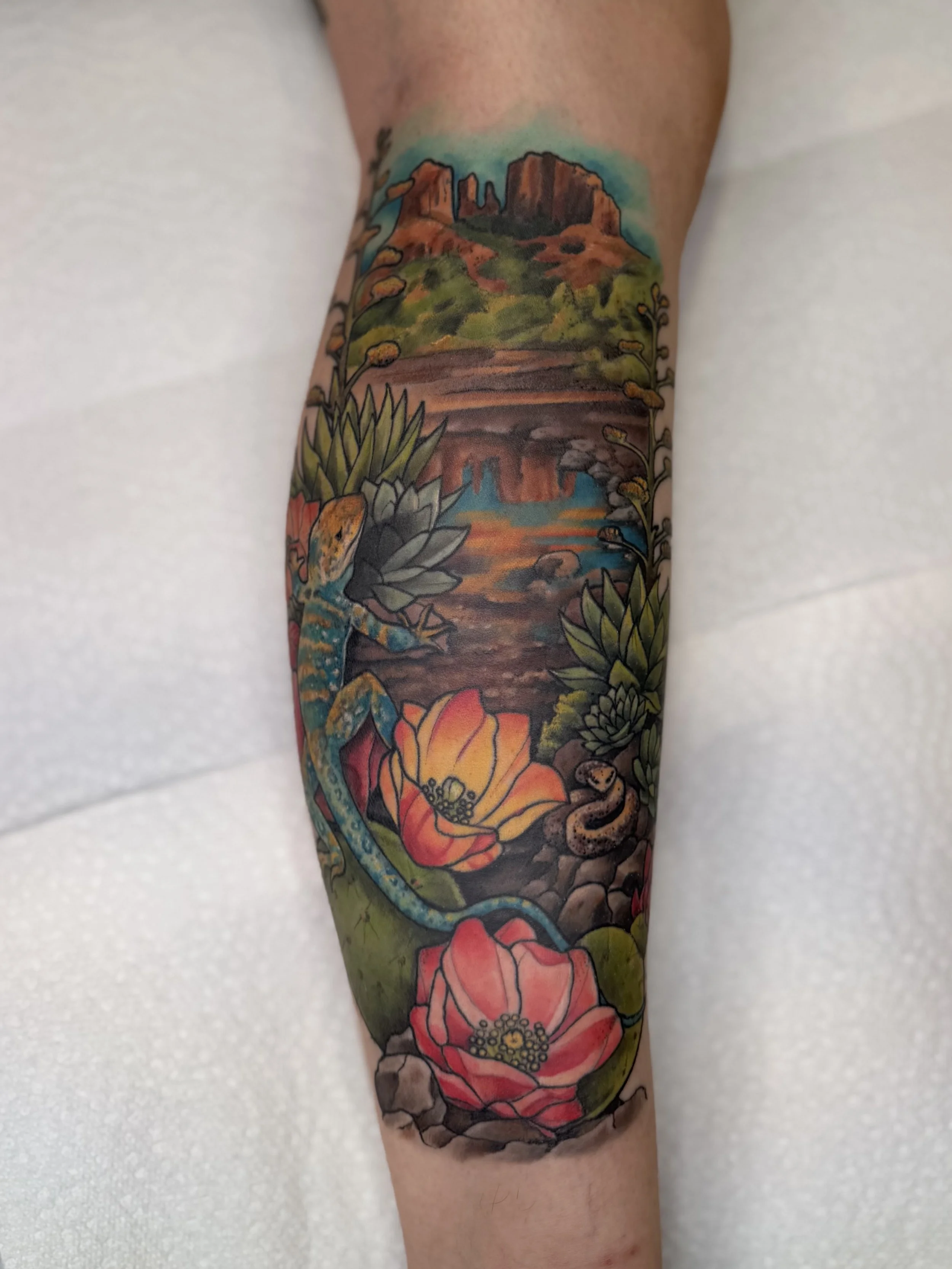 Colorful tattoo of a desert Sedona landscape with rock formations, a body of water, flowers, and plants on a person's arm. custom tattoo by Jordan Joyes  