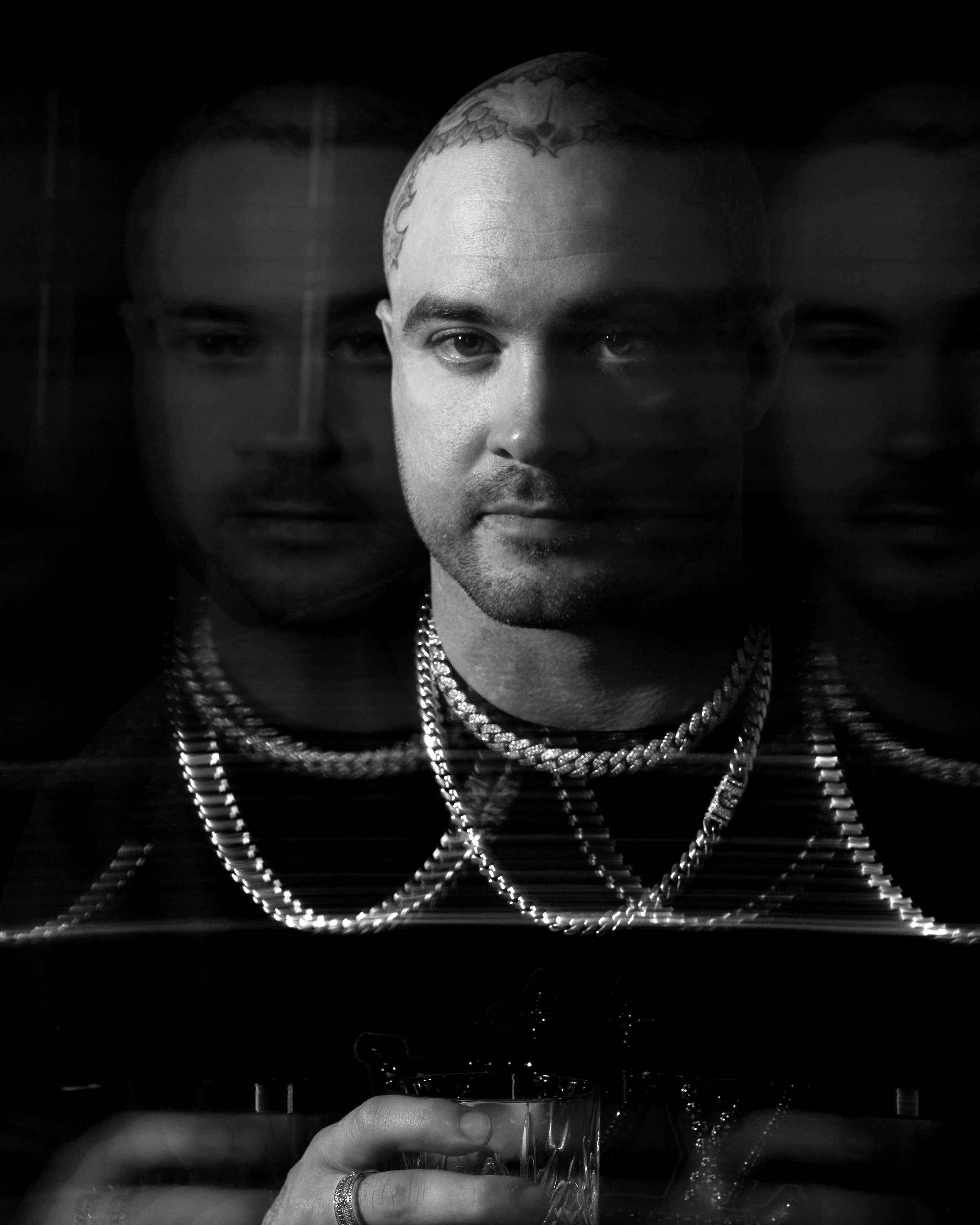 A black-and-white photo of a man with short hair and tattoos on his head, wearing multiple chains and holding a glass in his hand, with his reflection appearing twice behind him.