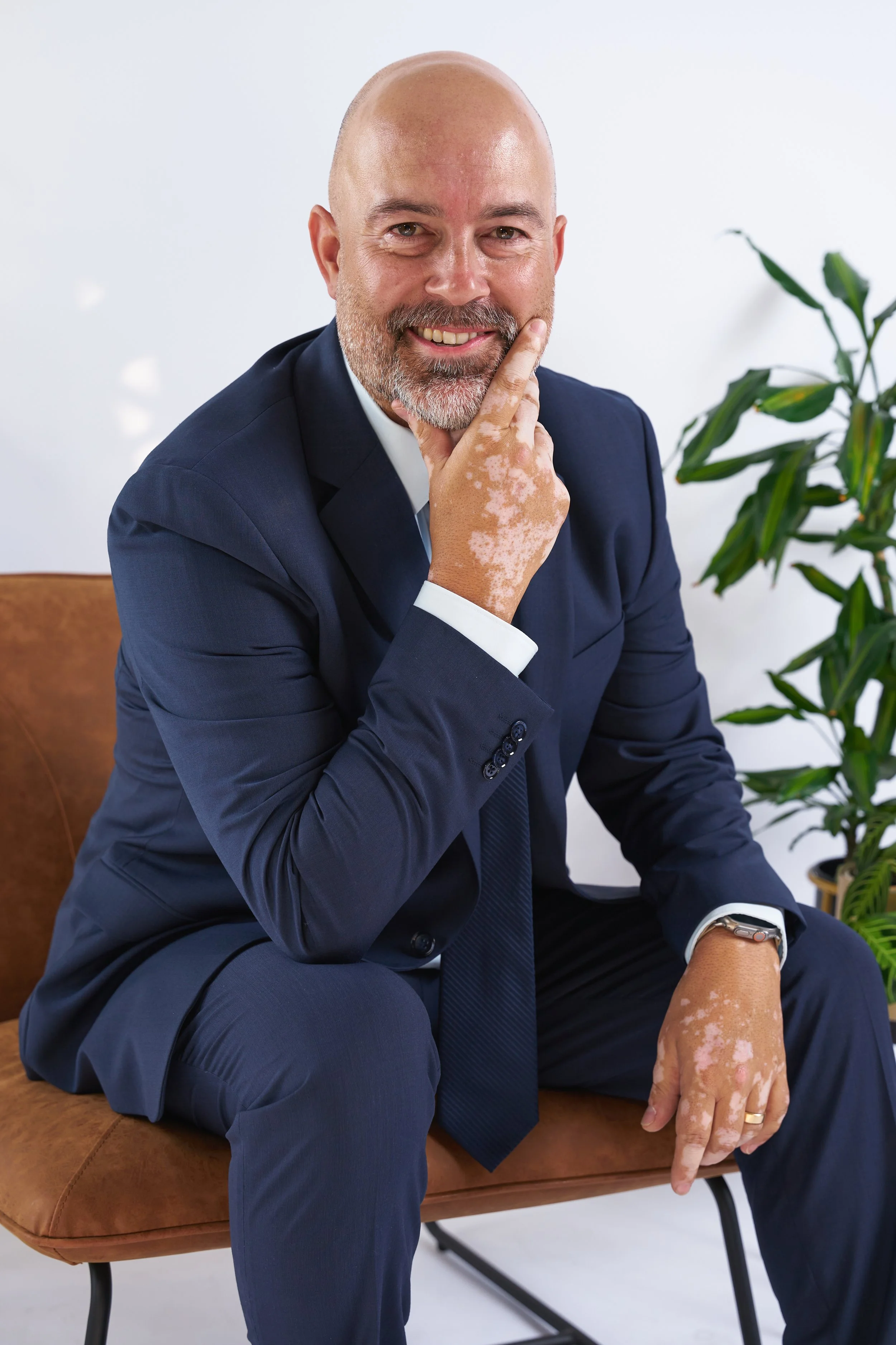 A bald man with a beard dressed in a navy suit sitting on a tan chair with a potted plant in the background. He is smiling, with one hand on his chin and the other resting on his knee.