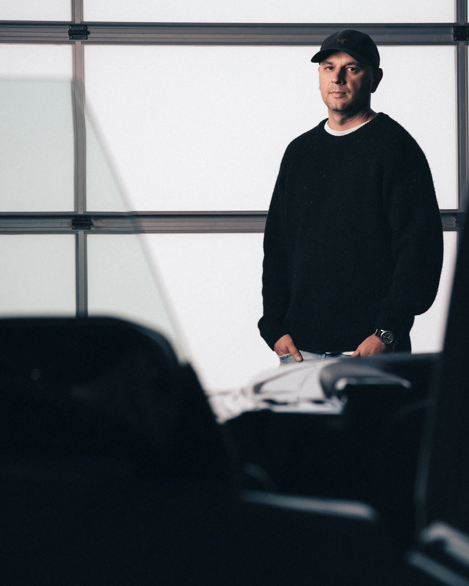 A man in a black sweater and cap standing in front of a white wall, partially obscured by a car in the foreground.