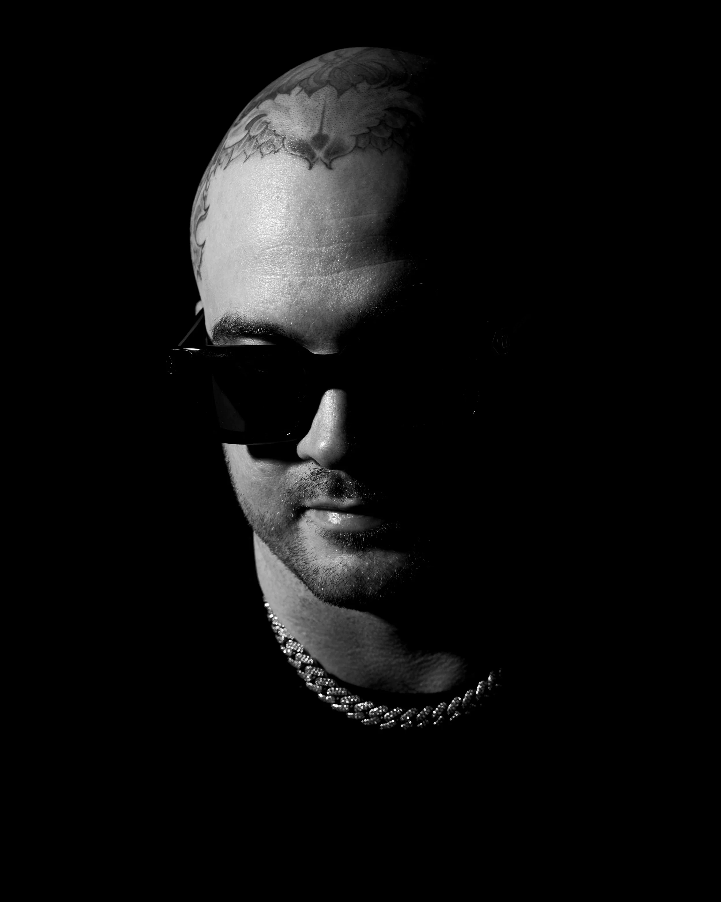 Black and white photo of a person with a shaved head, tattoo on the scalp, wearing sunglasses and a chain necklace, partially obscured by shadows.