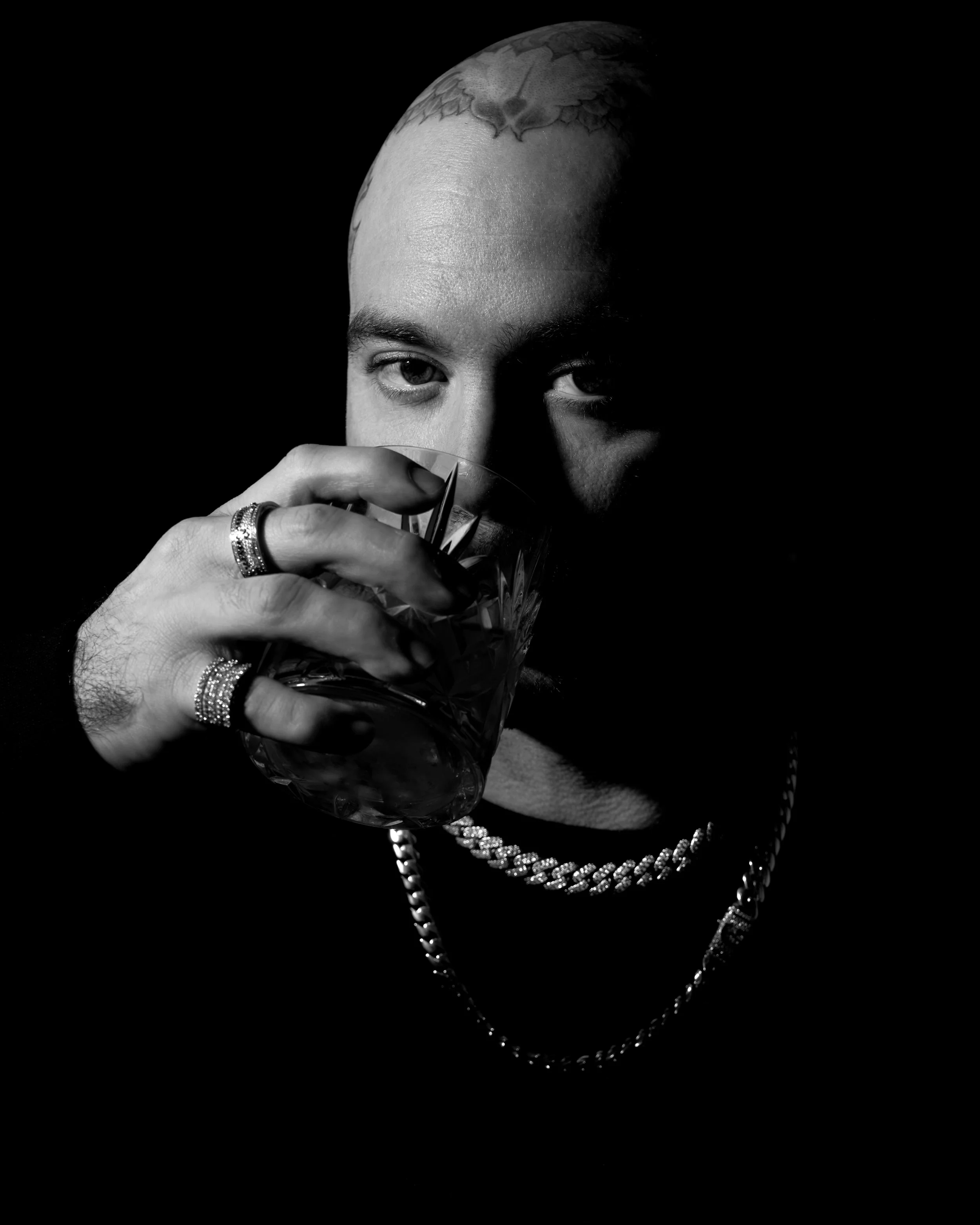 A person with tattooed head holding a glass of drink, wearing jewelry, with dark background