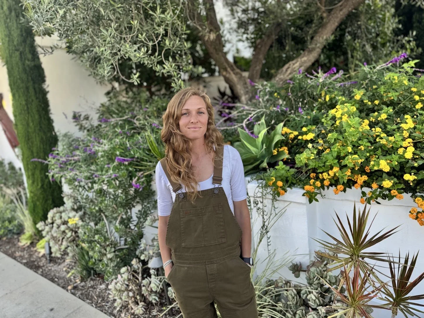A woman with wavy, shoulder-length blonde hair stands outdoors near a garden of colorful plants and flowers, with a large tree in the background.