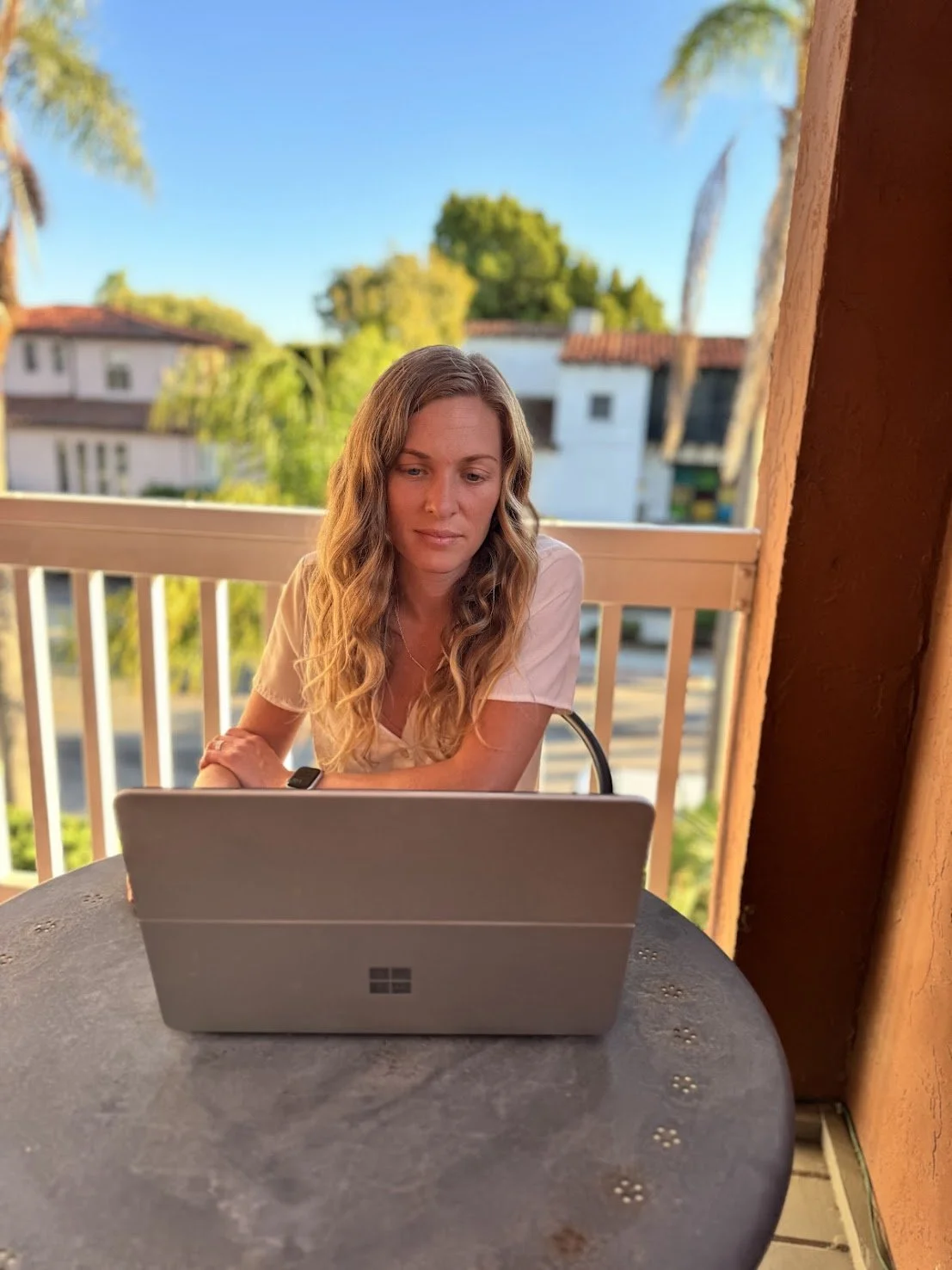 A woman with long wavy blonde hair sitting at a round outdoor table, looking at a Microsoft Surface laptop, with a balcony railing and trees in the background during sunset.