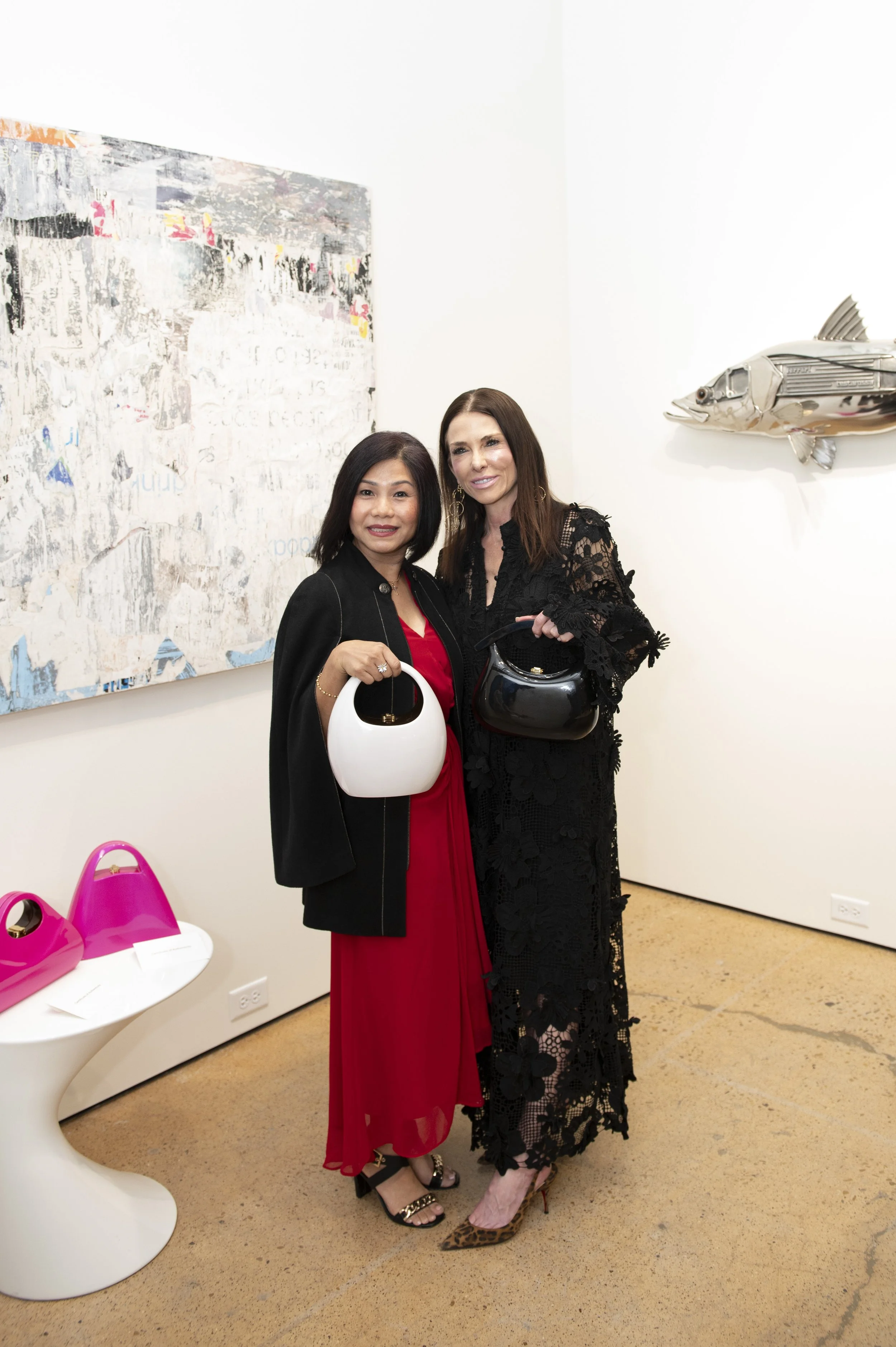 Two women at an art gallery, holding handbags and dressed elegantly, standing in front of abstract artwork and modern sculptures, including a fish-shaped sculpture on the wall.