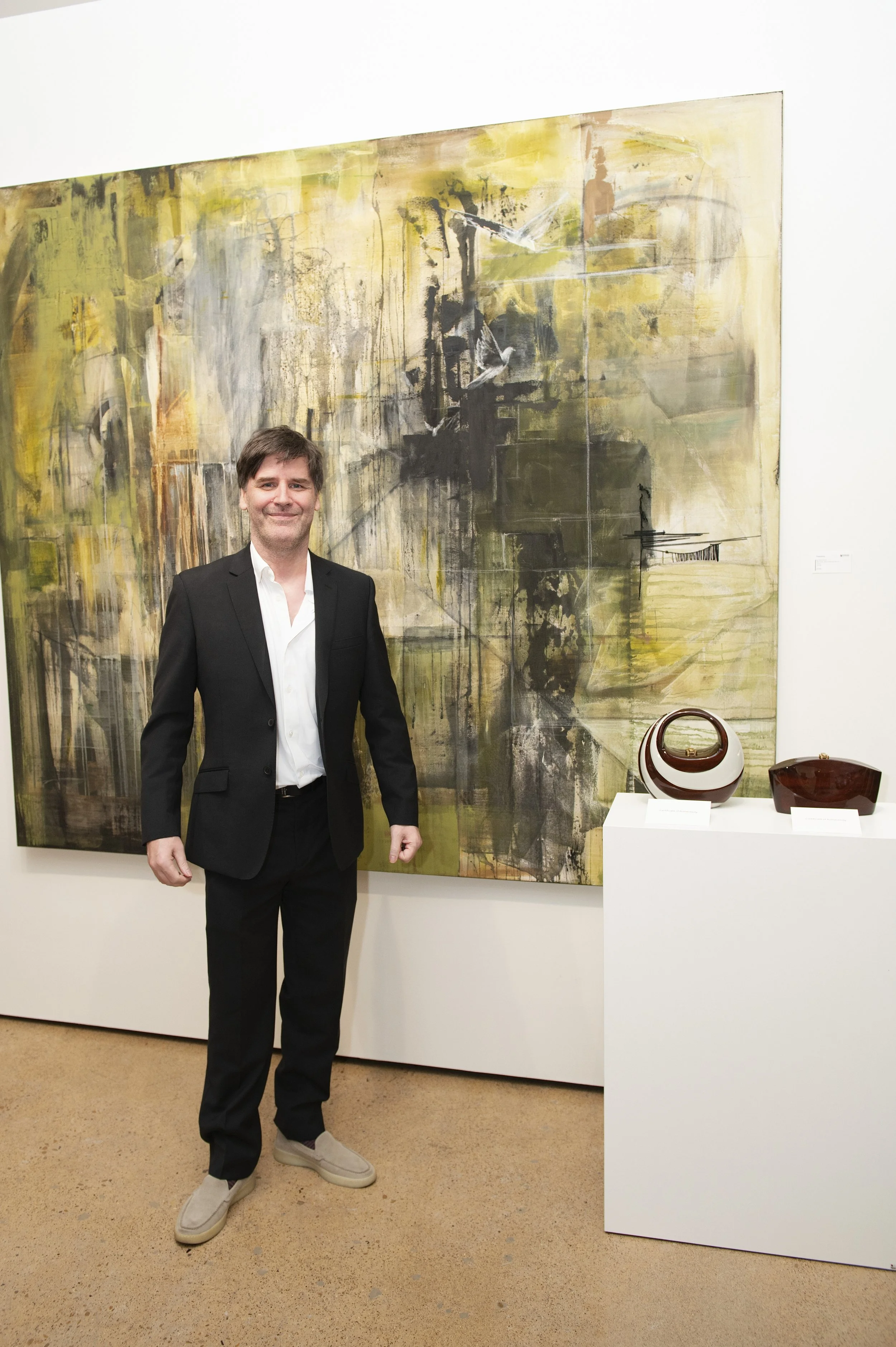 A man in a black suit and white shirt standing in an art gallery next to abstract paintings and pottery displays.