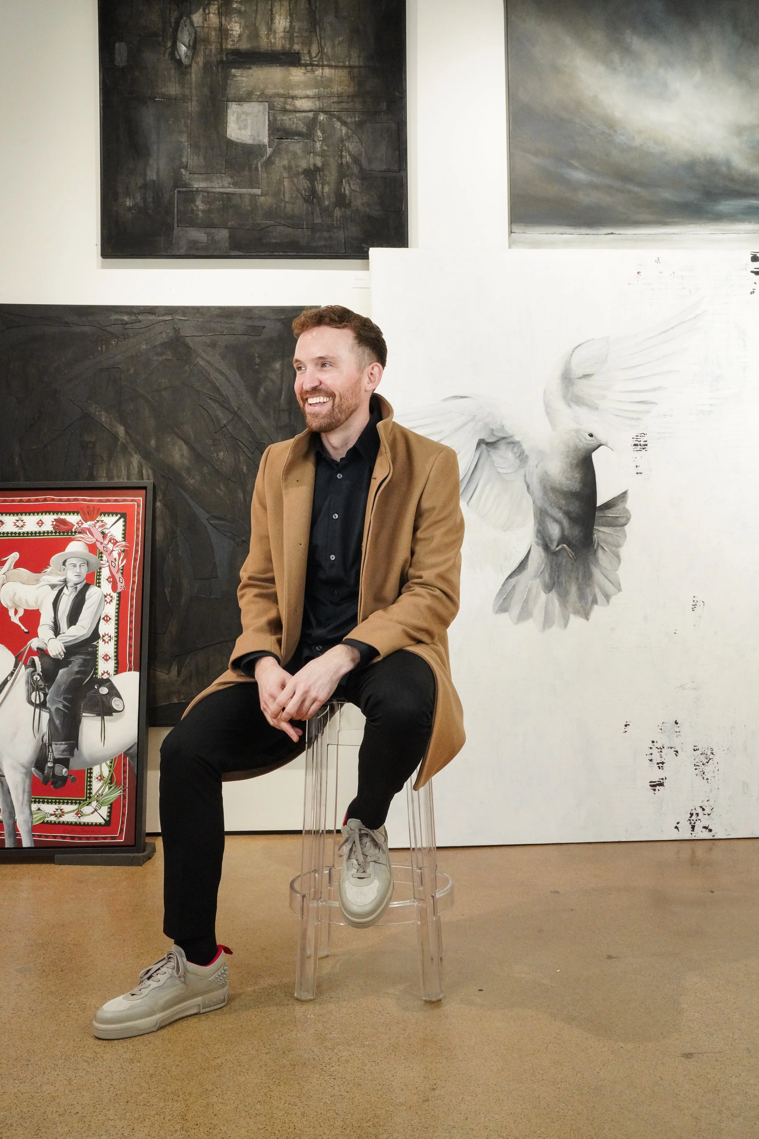 A man sitting on a transparent stool in an art gallery with abstract artwork and a painting of a man on a horse in the background.