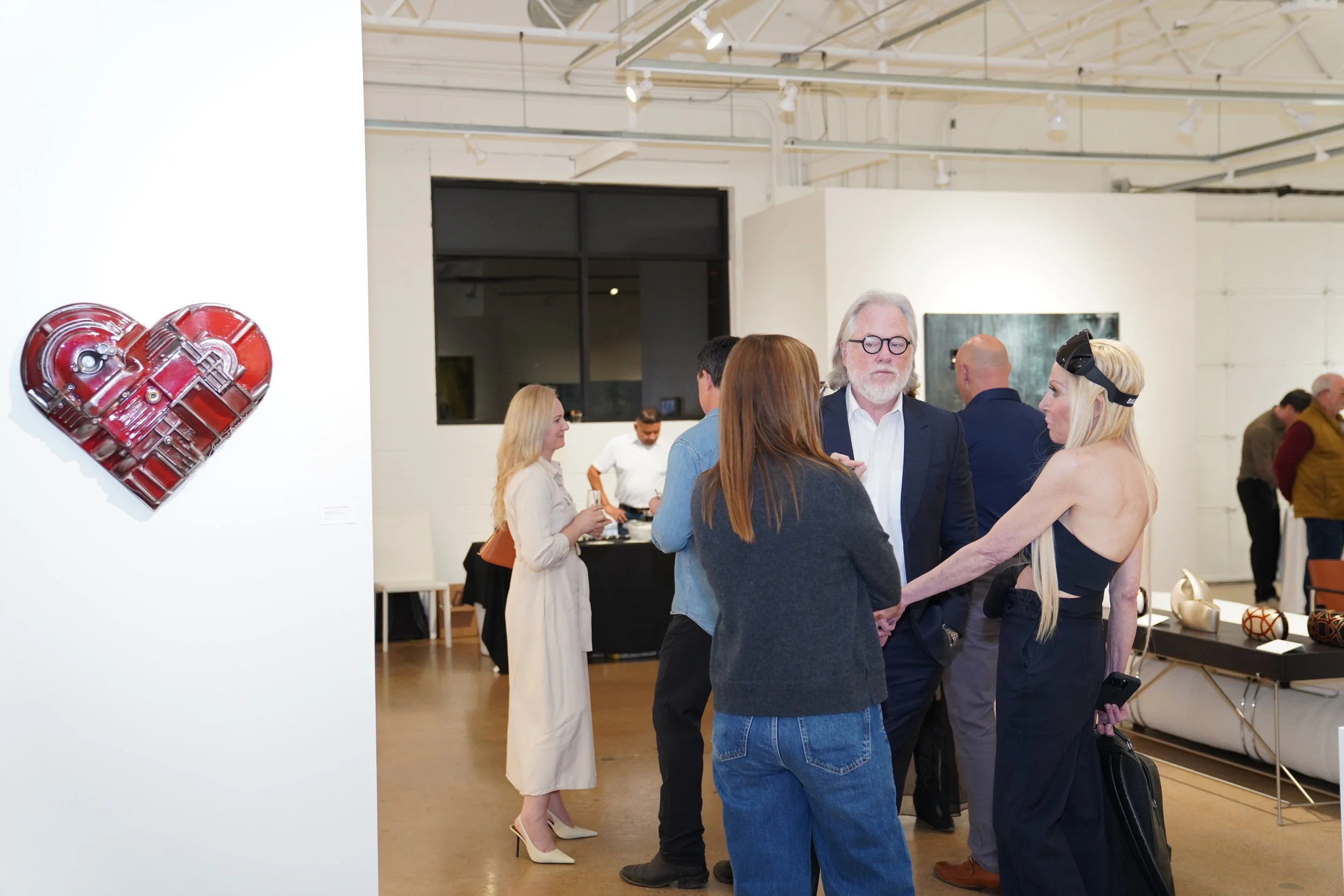 People socializing at an art gallery with a red heart-shaped sculpture on the wall.