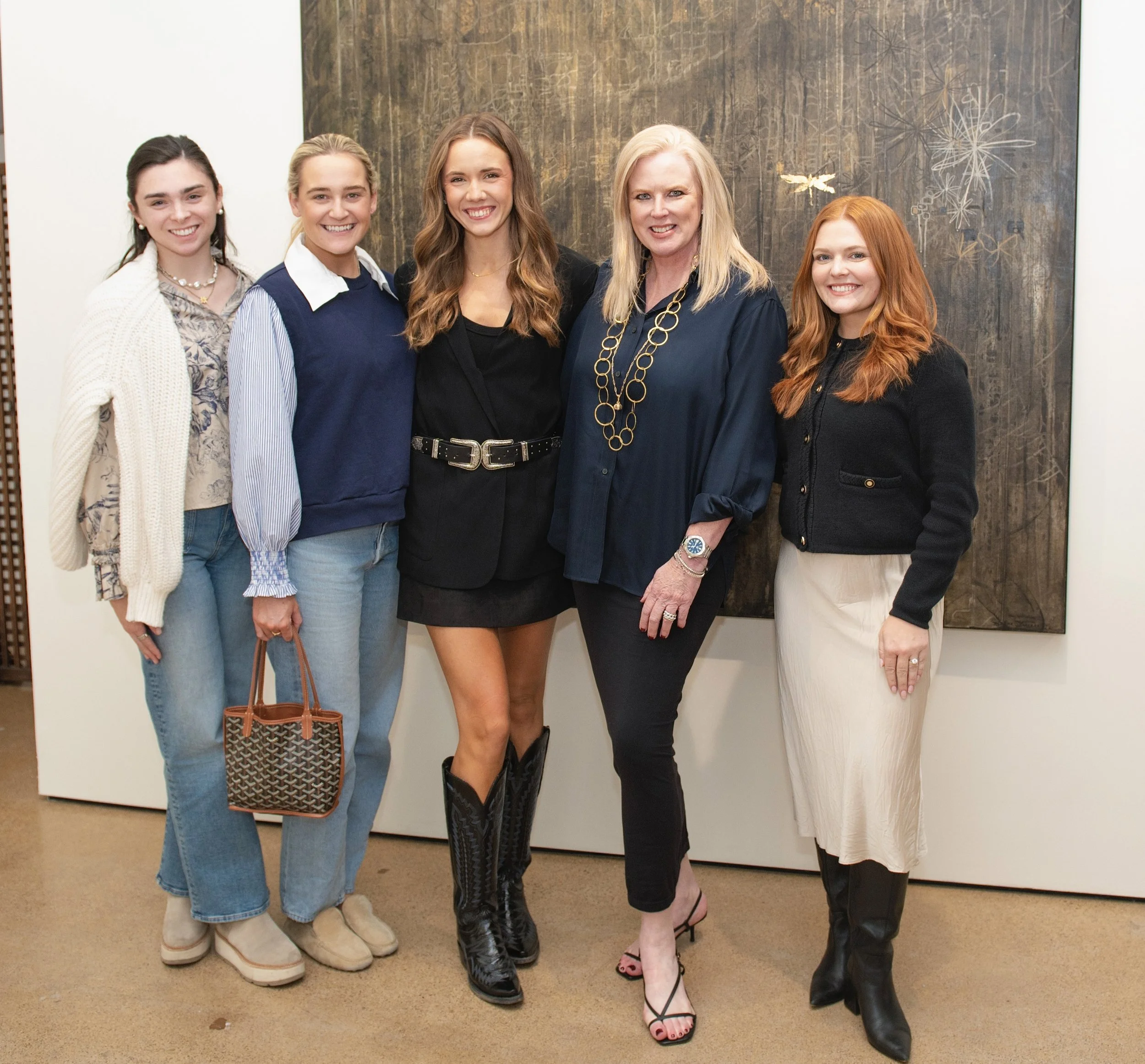 Five women standing together, dressed fashionably, smiling, in front of an art piece with firework and dragonfly designs.