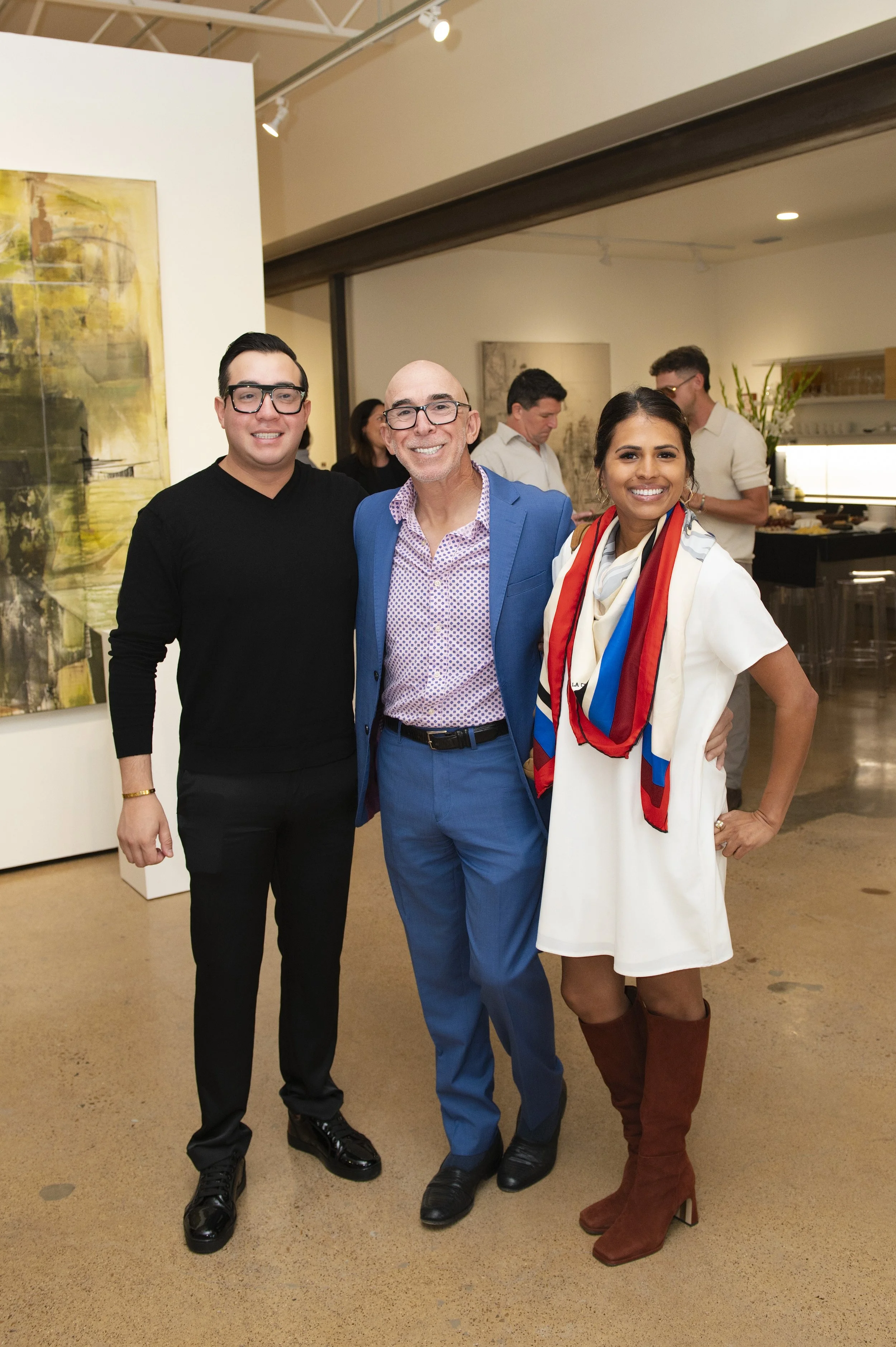 Three people smiling and posing together at an art gallery event. The man in the middle is wearing a blue suit and glasses. The woman on the right is wearing a white dress, brown knee-high boots, and a multi-colored scarf. The man on the left is dres