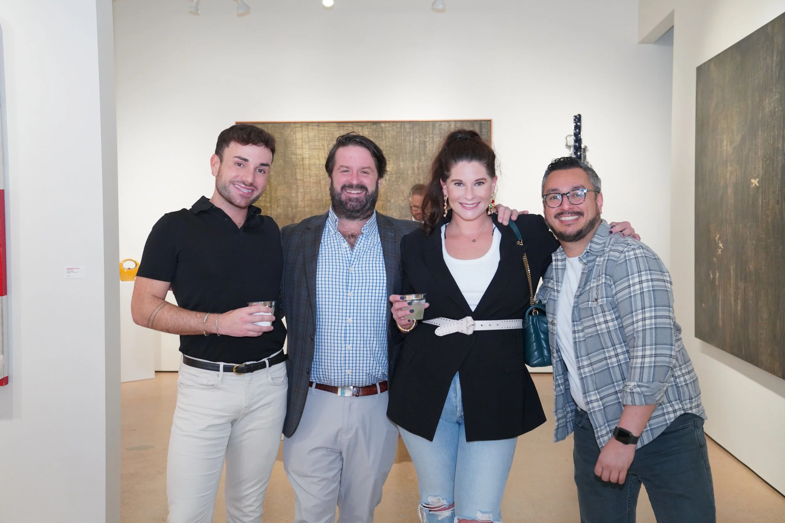Four friends standing together at an art gallery, smiling, with drinks in their hands. They are dressed casually and appear to be enjoying the event.