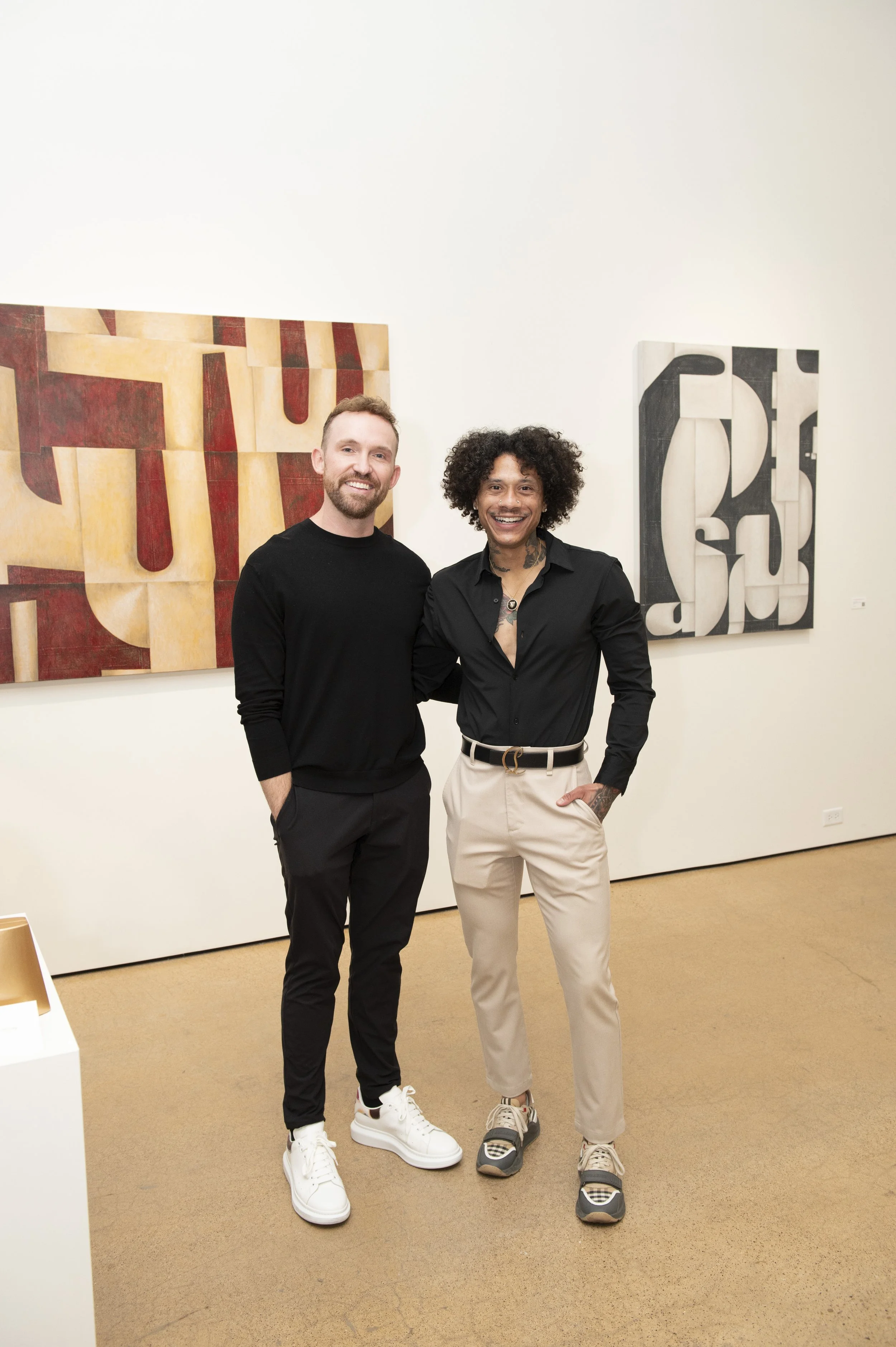 Two men standing together in an art gallery, smiling. The man on the left has light skin, a beard, and is wearing a black long-sleeve shirt and black pants. The man on the right has darker skin, curly hair, and is wearing a black shirt with beige pan