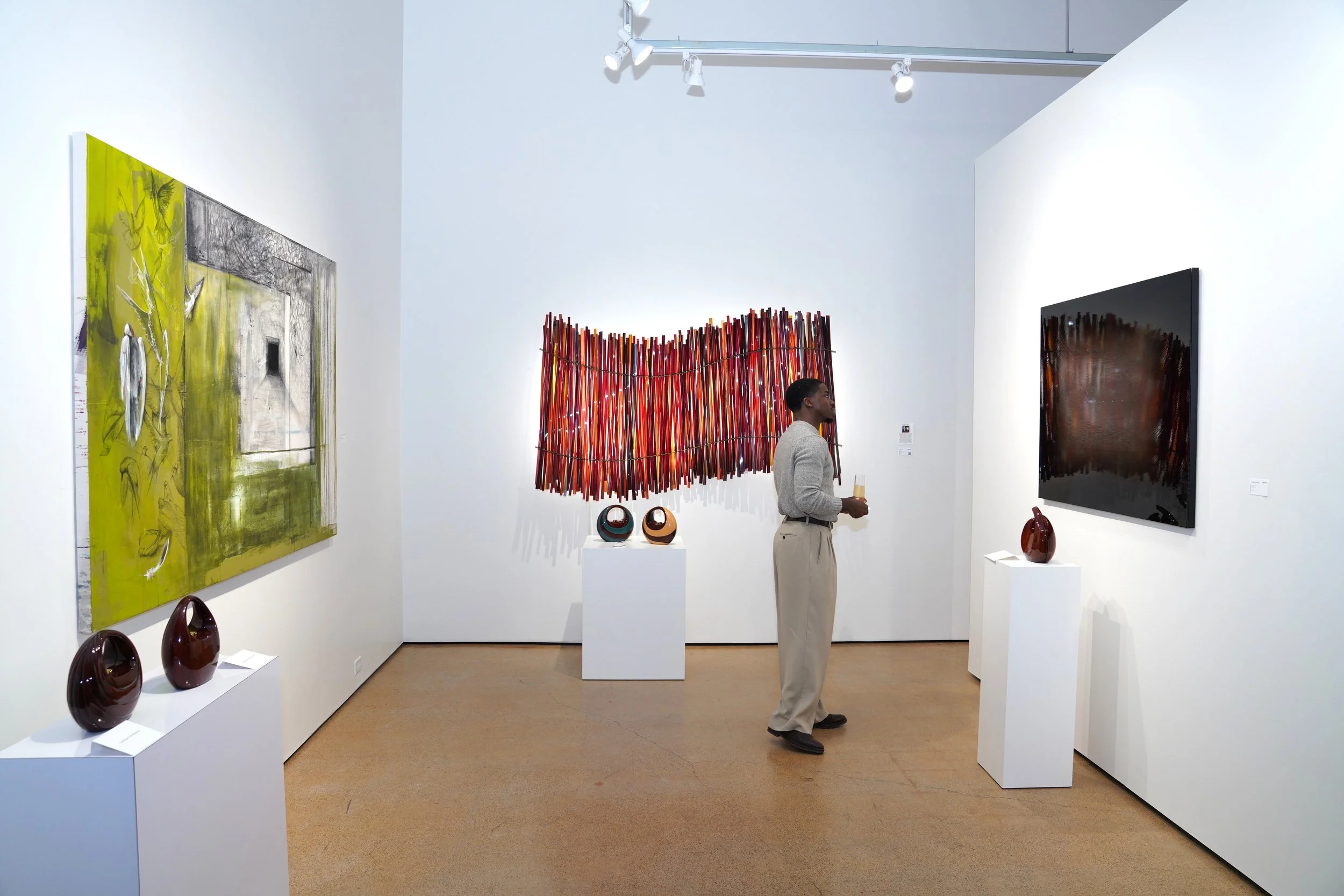 A man in a gray sweater and beige pants looking at abstract art in a gallery, surrounded by painted and sculpted art pieces on white pedestals and walls.
