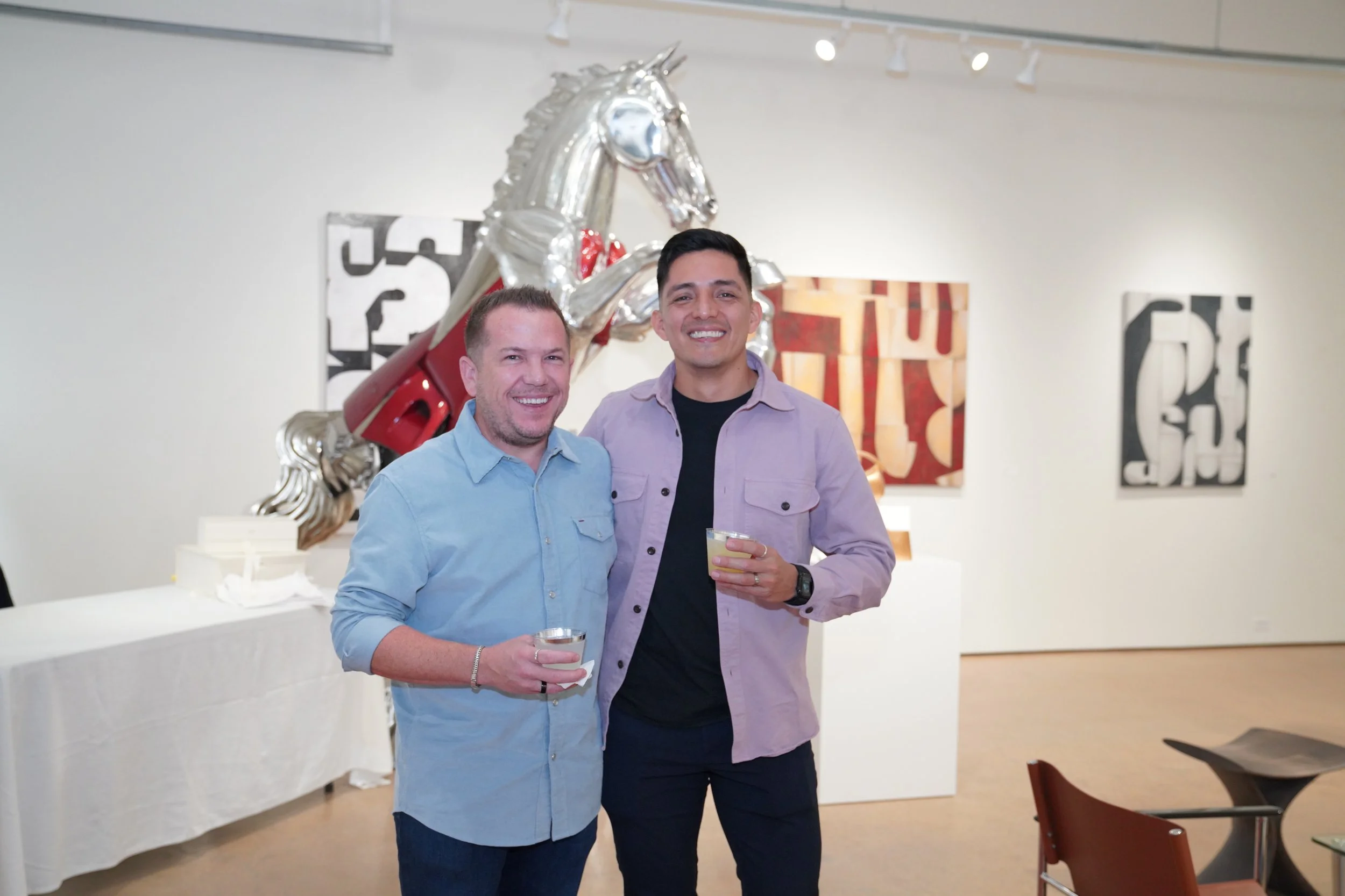 Two men holding drinks and smiling at an art gallery, with a large metallic unicorn sculpture and contemporary paintings on the wall behind them.
