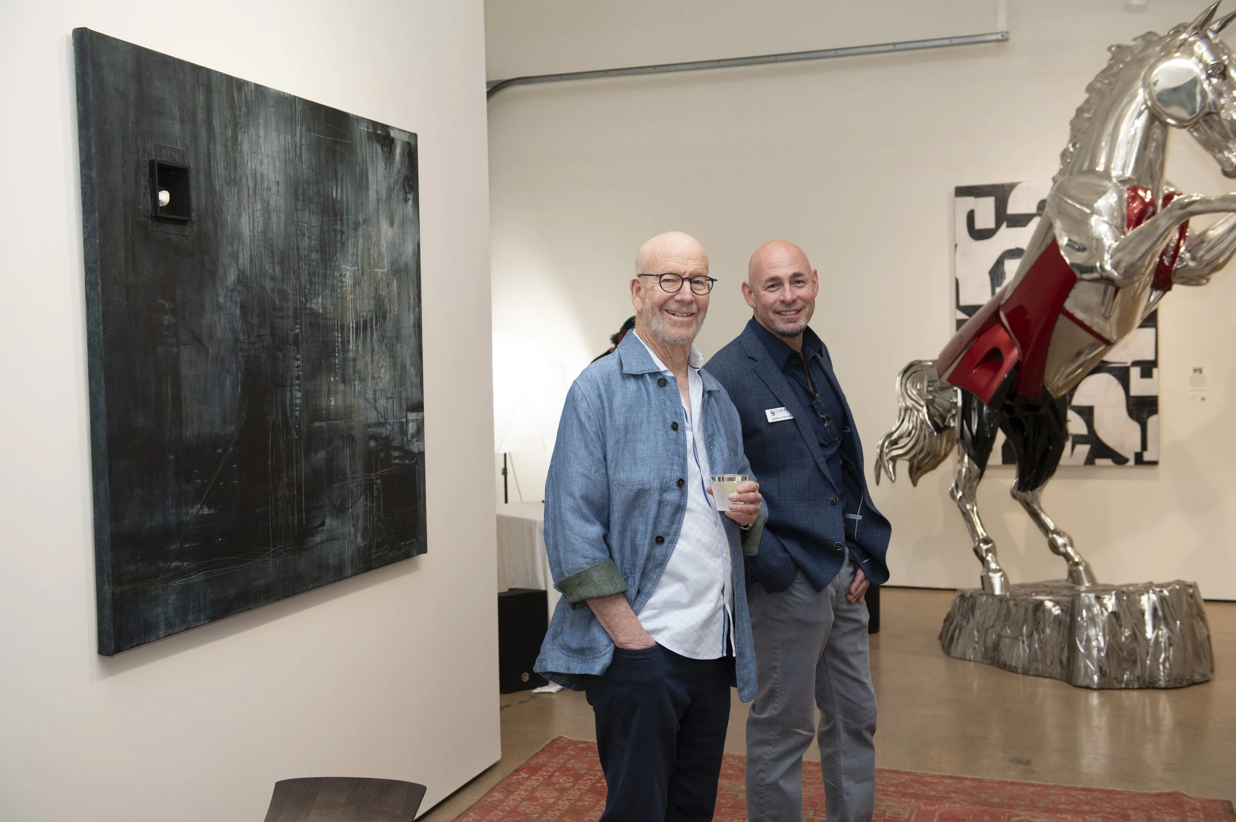 Two men smiling and standing in an art gallery, one holding a glass of water. Behind them, there is a modern abstract painting on the wall and a large metallic horse sculpture.