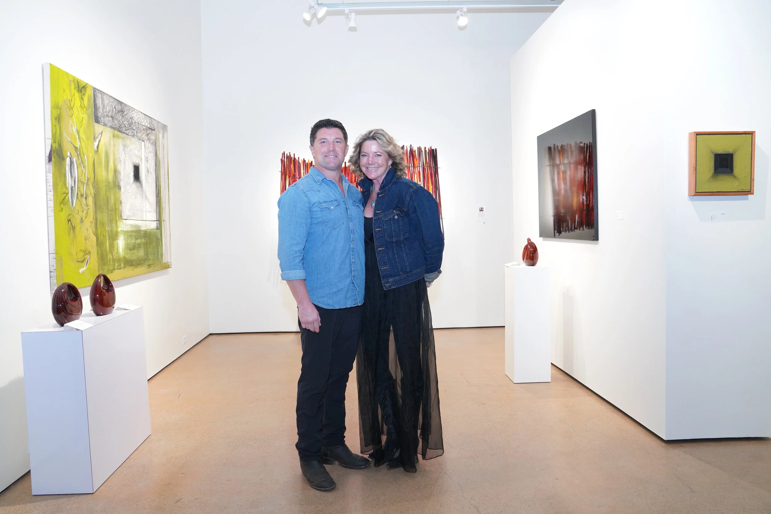 A man and woman smiling at an art gallery, standing close together surrounded by abstract paintings and sculptures on white walls and pedestals.
