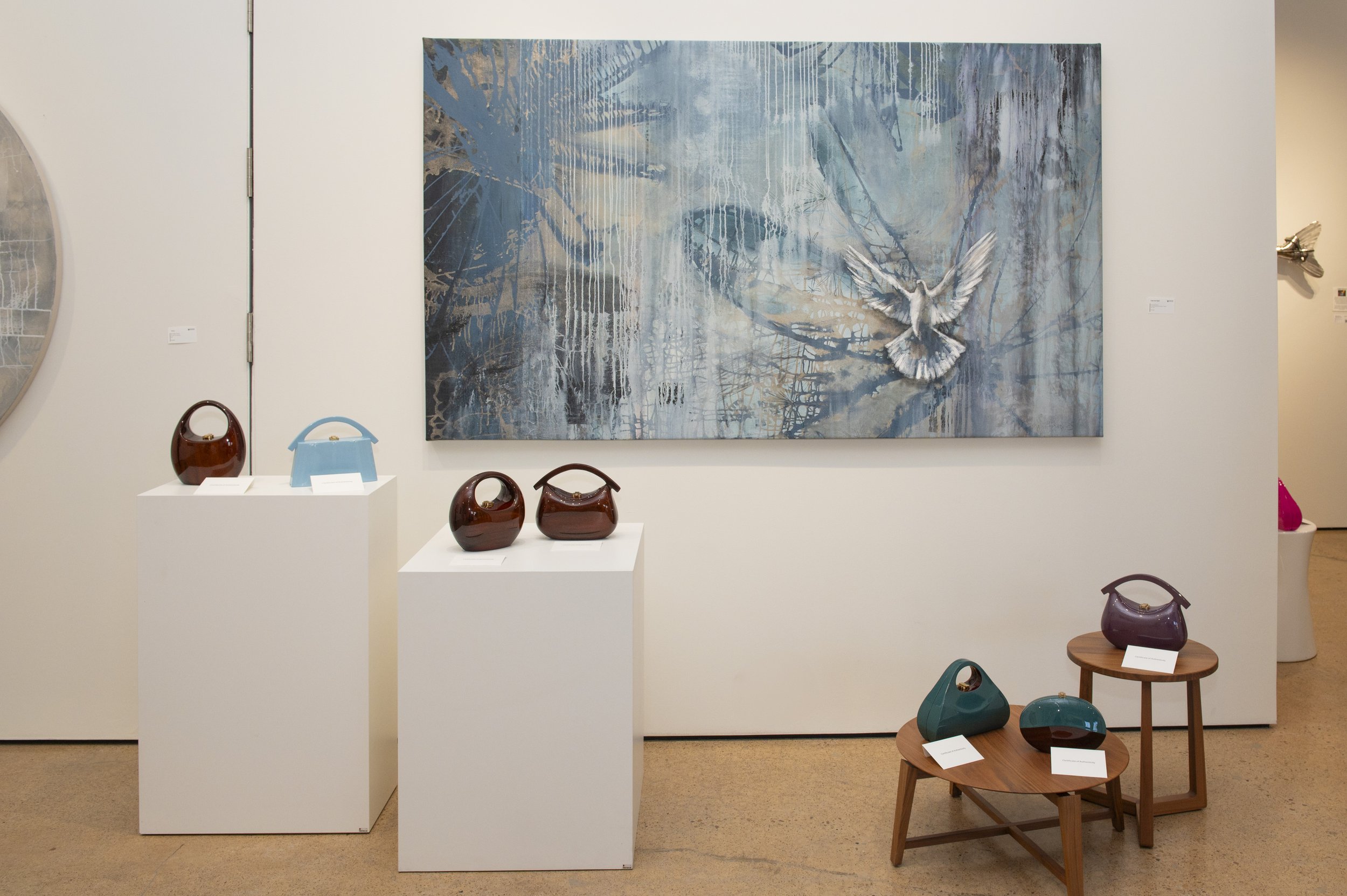 An art gallery display with a large abstract painting of blue and white tones. Below it, three polished ceramic vases in shades of brown and purple on white pedestals. To the right, two small wooden tables hold colorful ceramic sculptures, including 