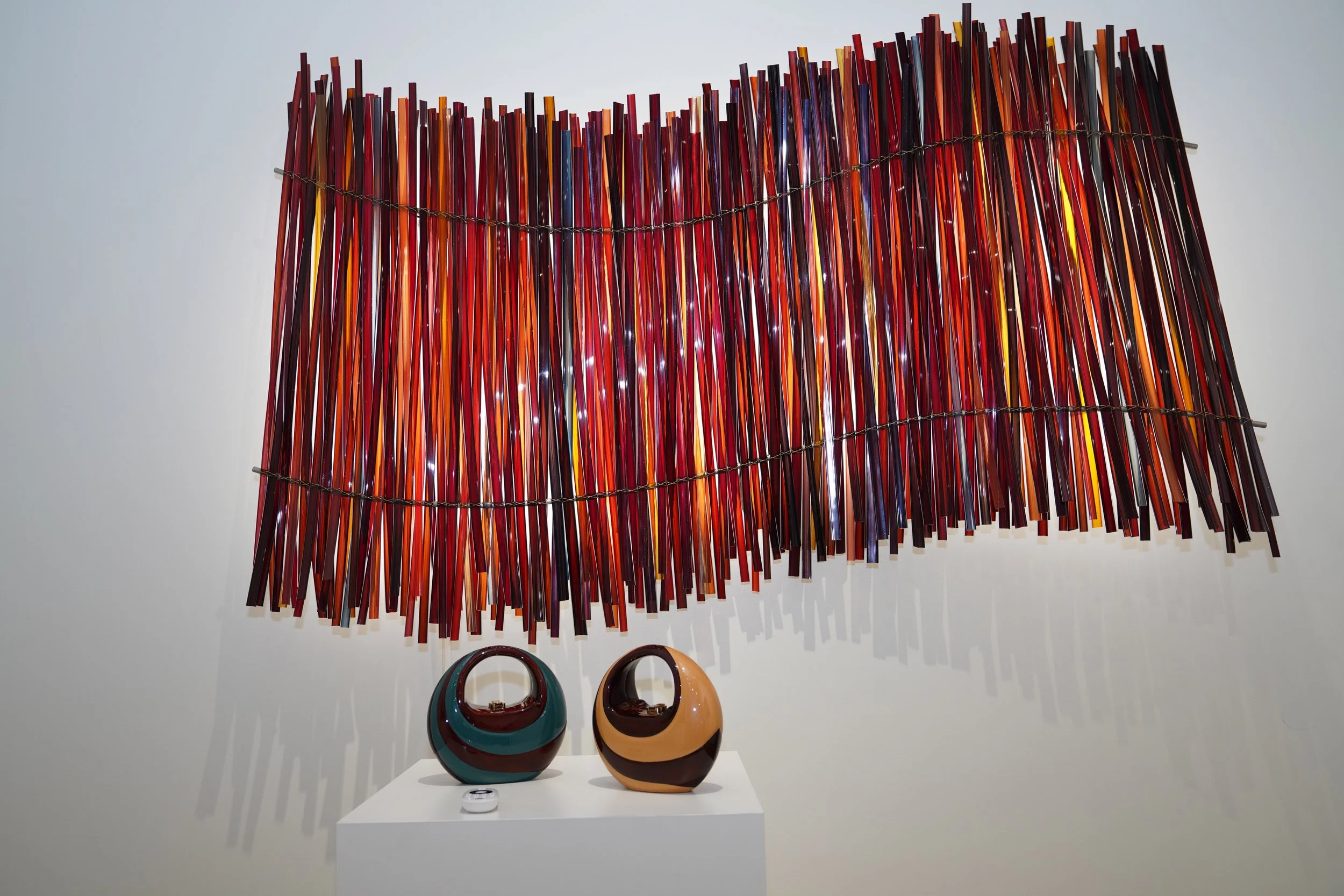 An art installation featuring a large wall hanging made of numerous colorful, elongated glass pieces in red, orange, yellow, and dark hues, arranged in a wavy pattern. In front of the wall are two decorative vases with rounded bodies and handles, one