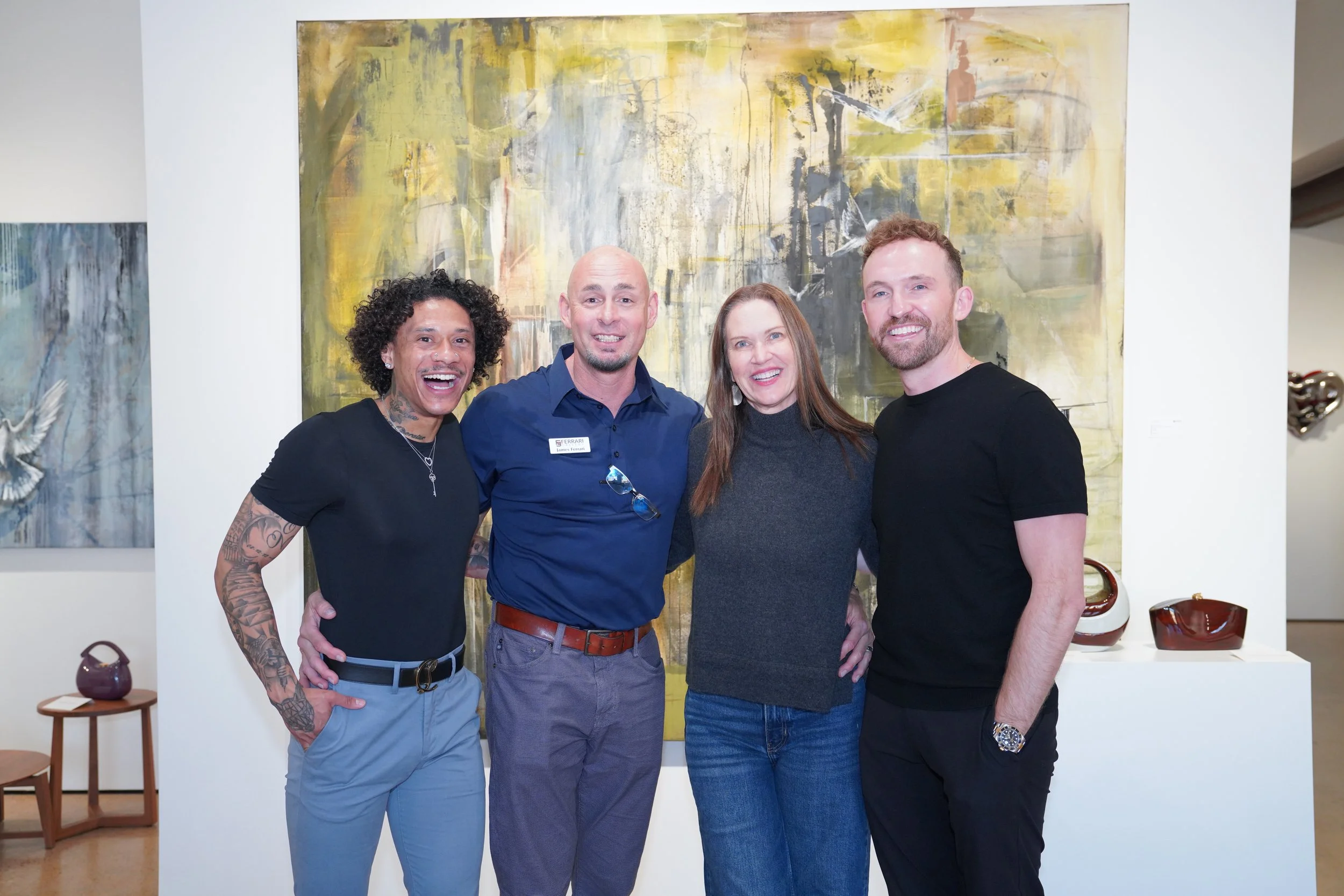 Four smiling people standing together in an art gallery with abstract paintings in the background, three men and one woman, dressed casually.