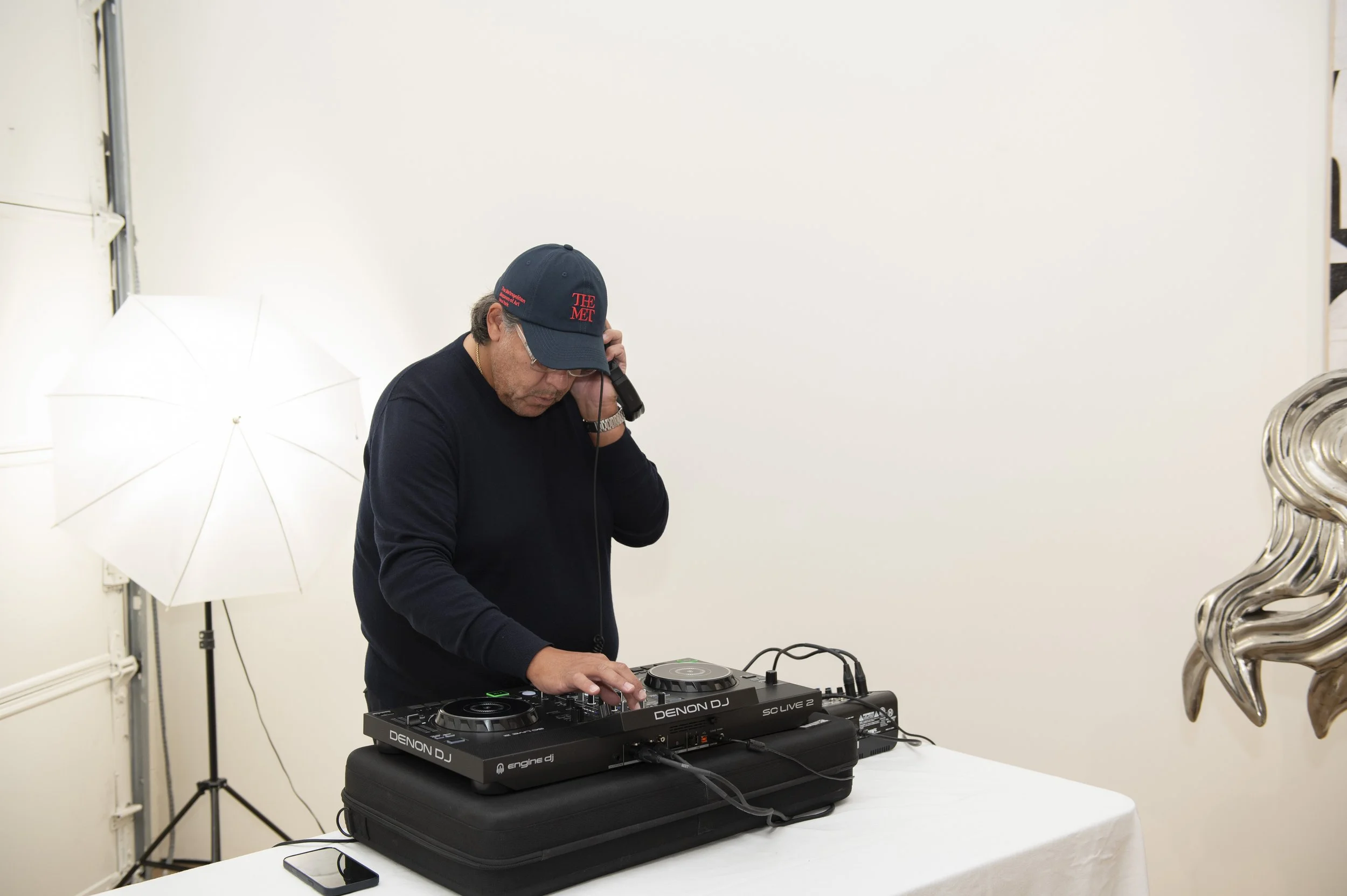 DJ wearing a cap and glasses, using DJ equipment at a white table, with a white umbrella light and a metallic sculpture in the background.