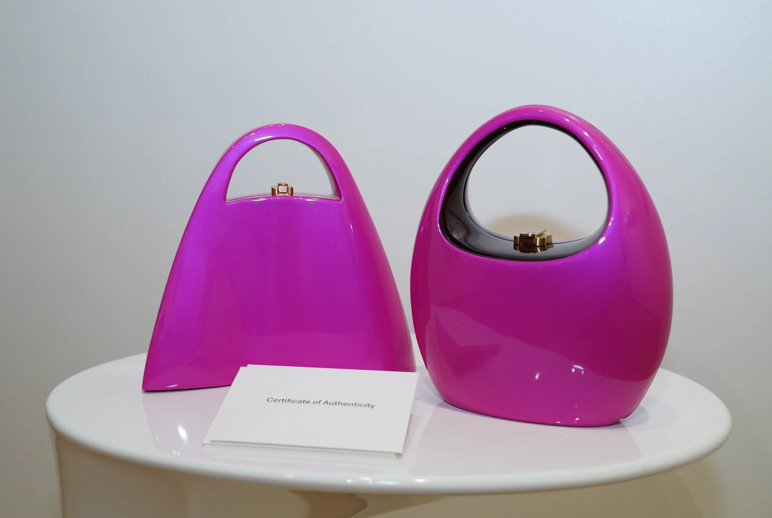 Two pink handbags with curved handles on a white table, one with a glossy finish and one with a cutout handle, accompanied by a Certificate of Authenticity.
