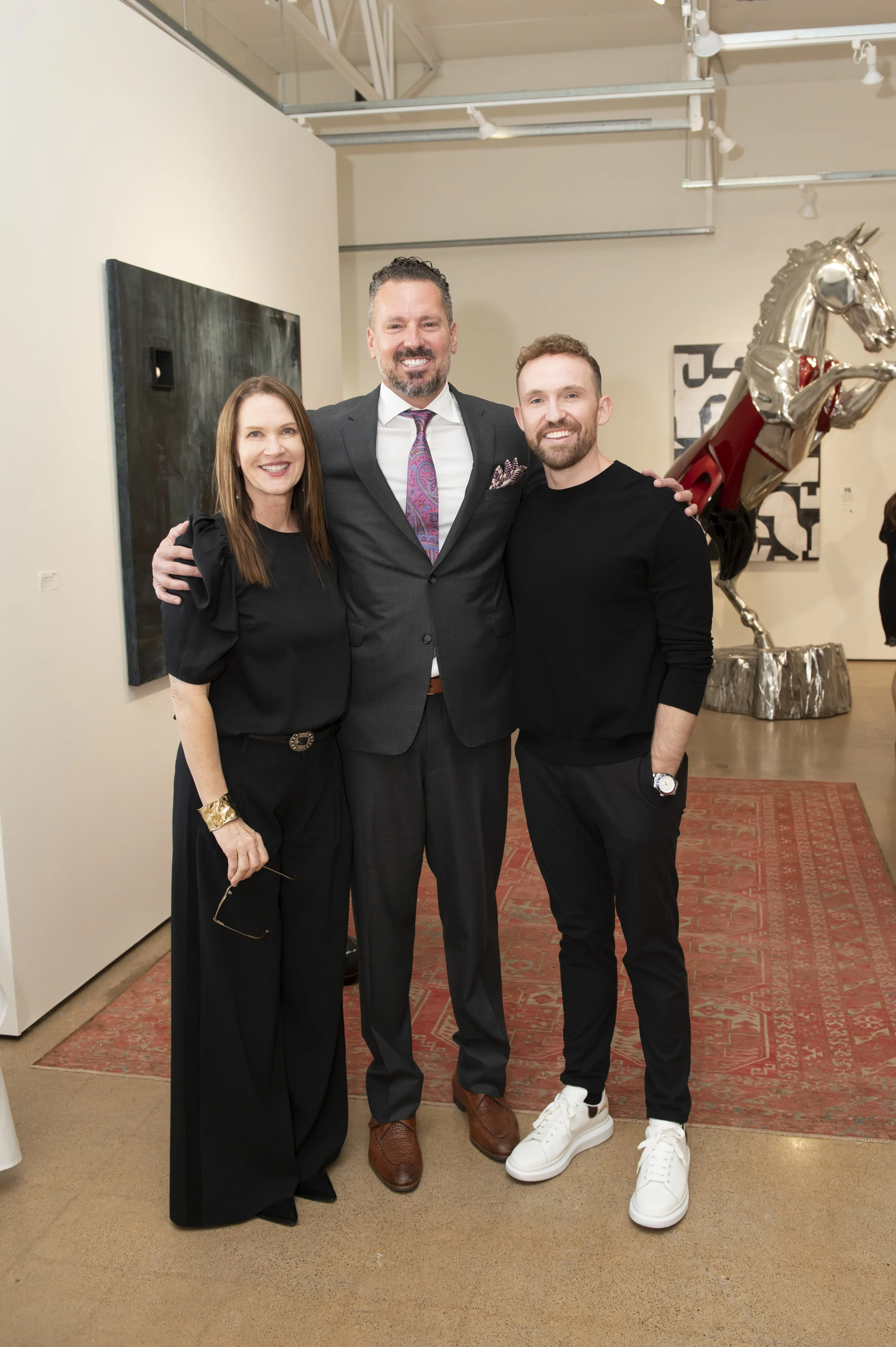 Three people standing together at an art gallery, smiling for the photo. The person on the left is a woman with long brown hair, wearing a black top with puffed sleeves and black pants. The person in the middle is a man with short dark hair and a bea