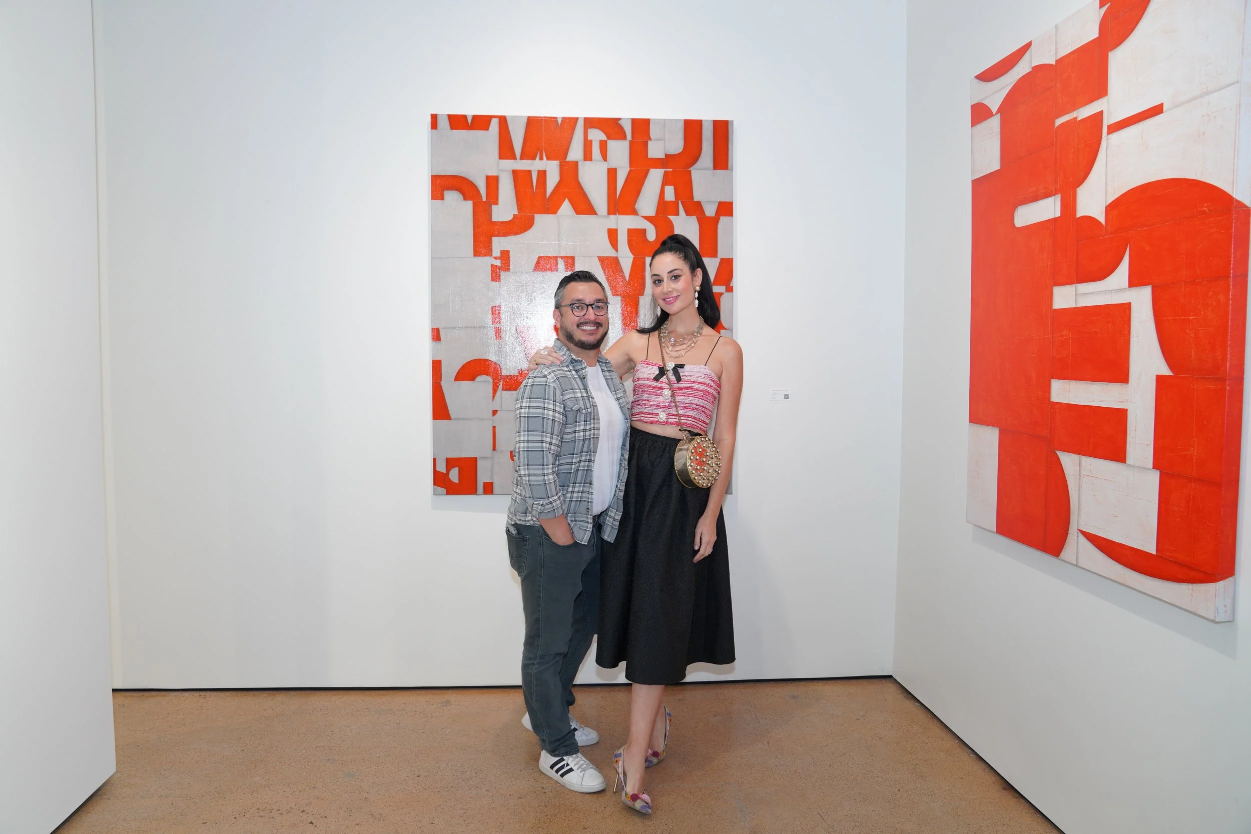 A man and woman standing together at an art gallery exhibition, smiling at the camera. The man has glasses, a beard, and is wearing a plaid shirt with jeans. The woman has long black hair, jewelry, a colorful strapless top, a black midi skirt, and co