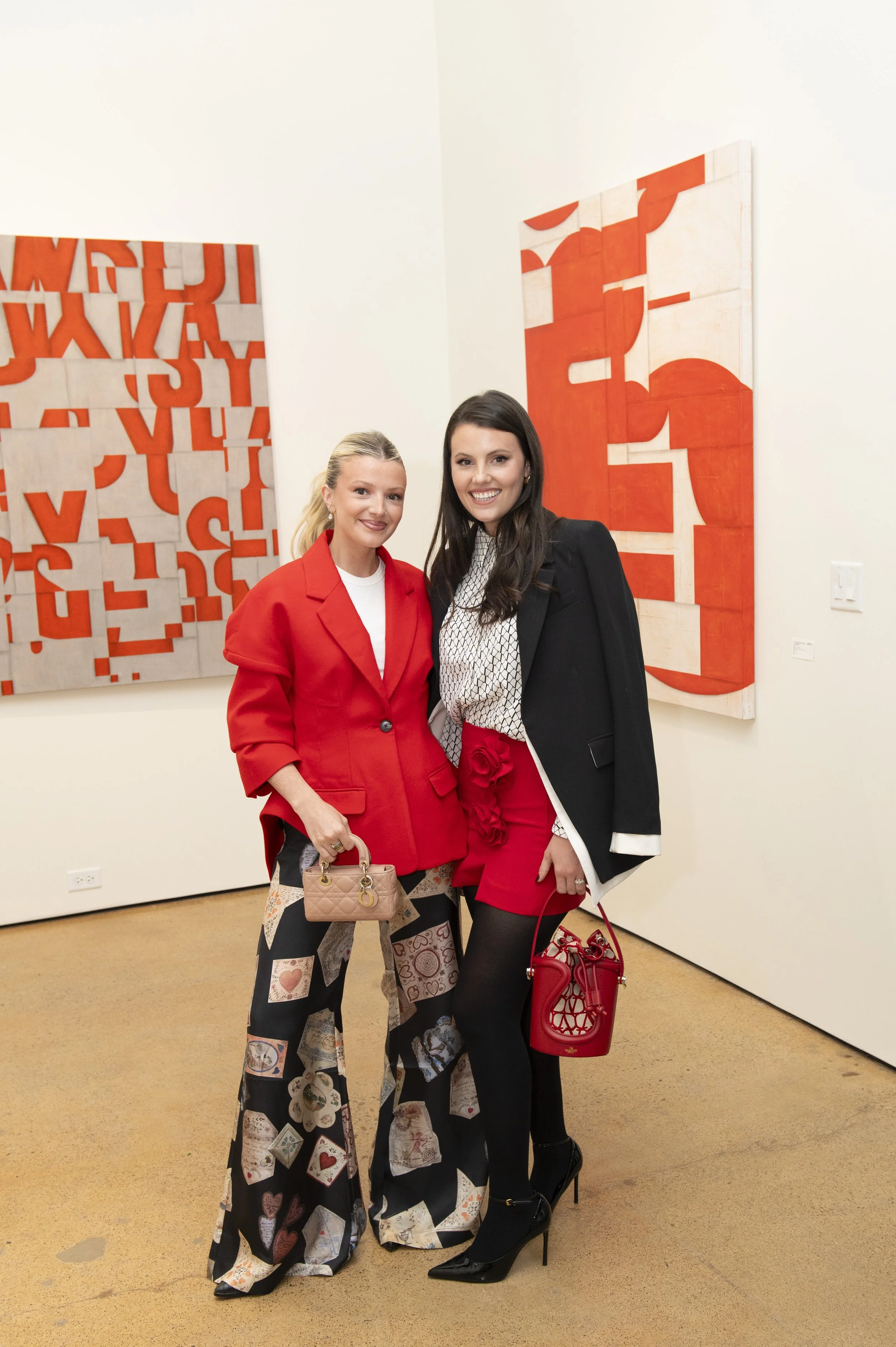 Two women standing together at an art gallery, smiling, with abstract red and white paintings behind them. One woman wears a red blazer and patterned wide-leg pants, holding a small beige handbag. The other woman wears a black blazer over a patterned
