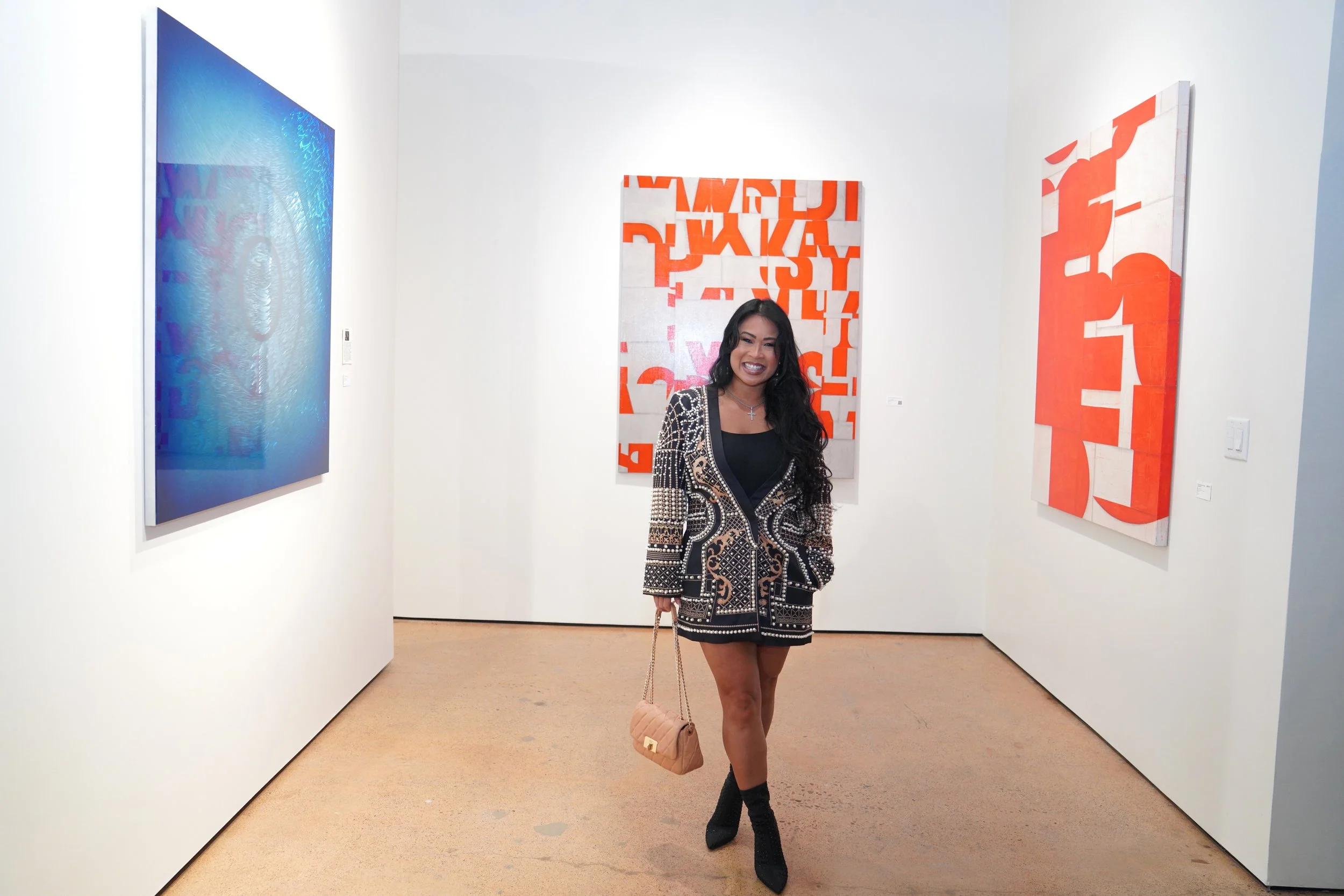 A woman with long dark hair smiling in an art gallery, wearing a patterned blazer, black top, and black boots, holding a beige purse, surrounded by abstract colorful paintings on white walls.