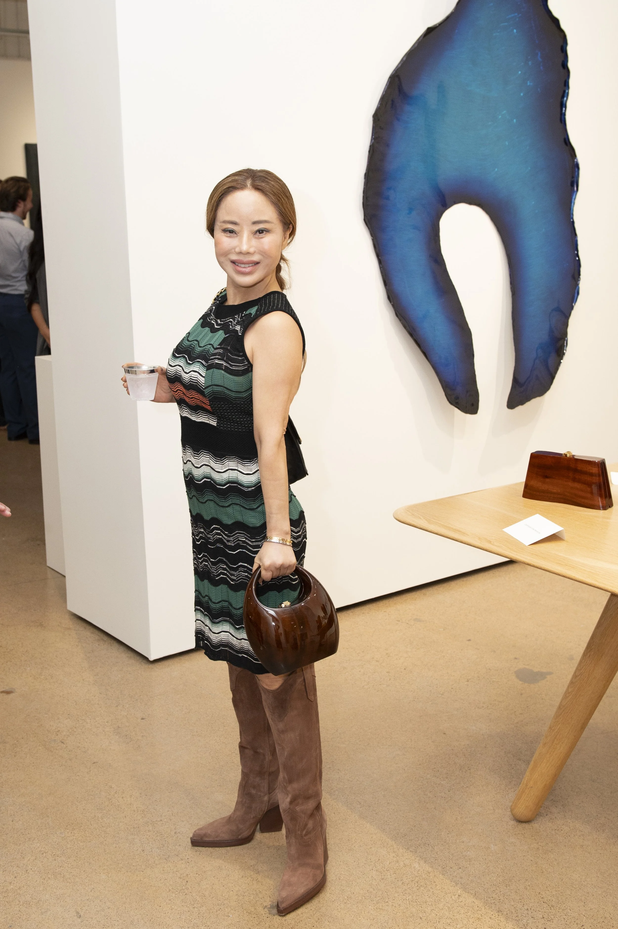 A woman standing at an art gallery, holding a drink and a brown purse, smiling at the camera. She is wearing a striped dress, tan boots, and has brown hair. Behind her is a large, abstract blue artwork on the wall and a wooden table.