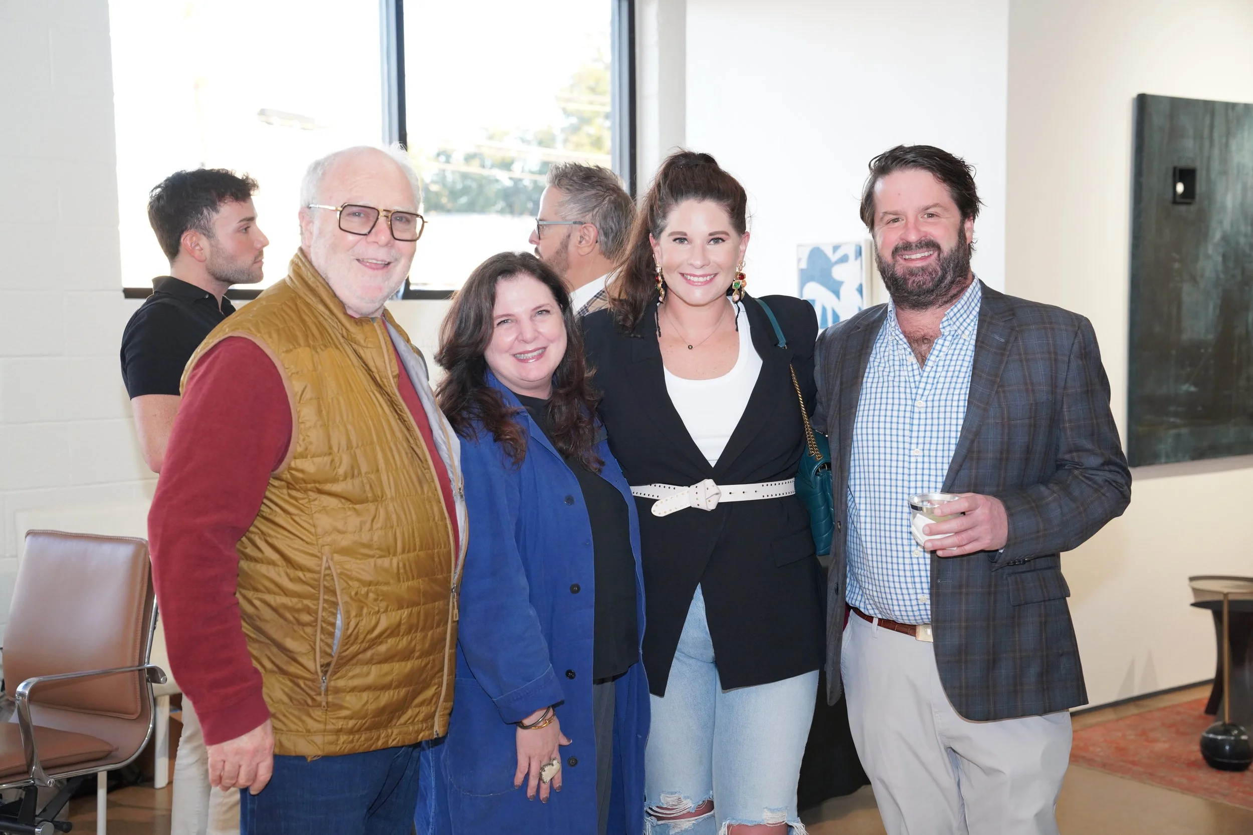 Group of five people smiling at an indoor social event, four standing in the foreground and one in the background, with artwork on white walls and a window behind them.