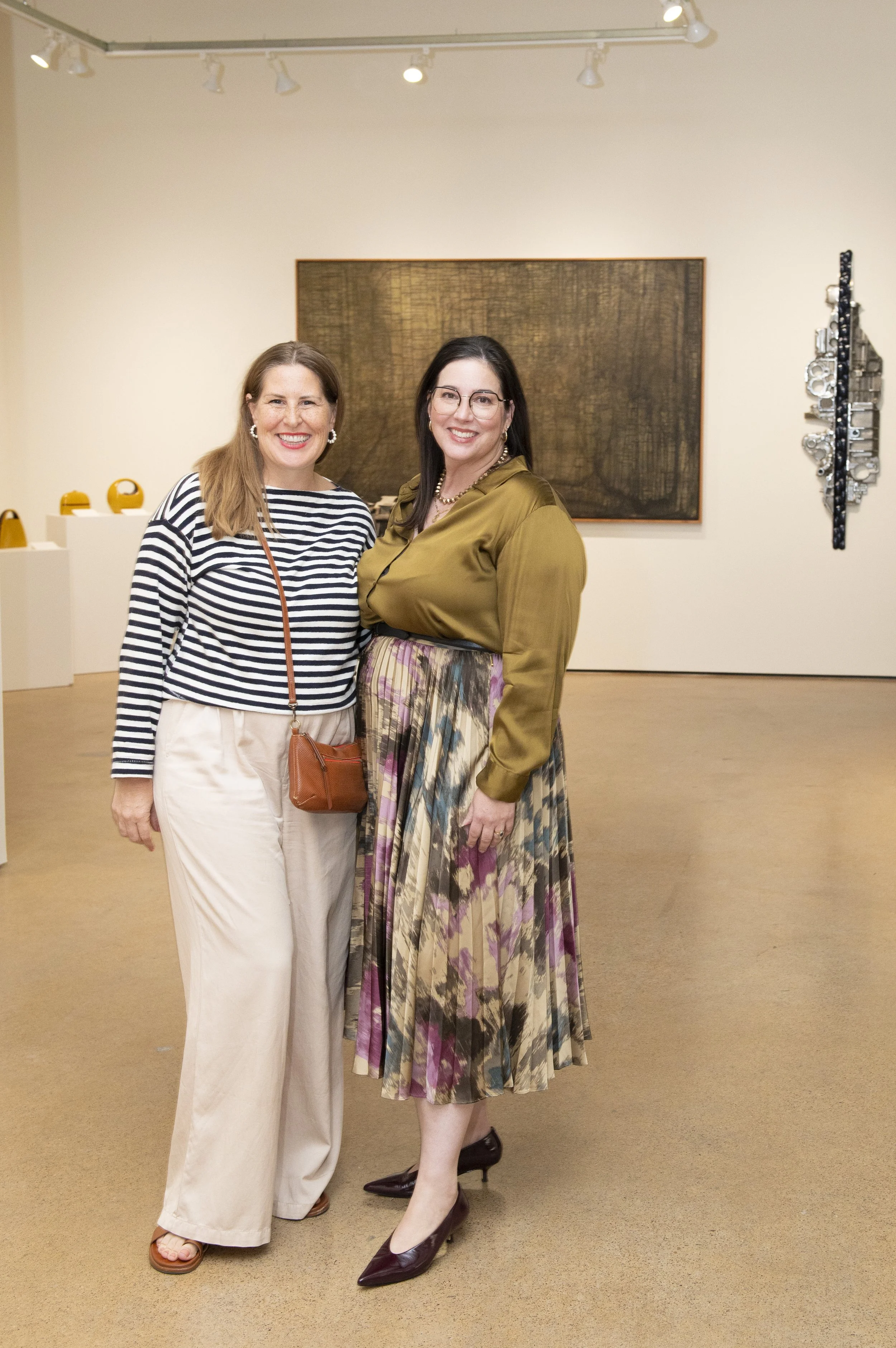 Two women standing and smiling in an art gallery with artworks on the wall behind them.
