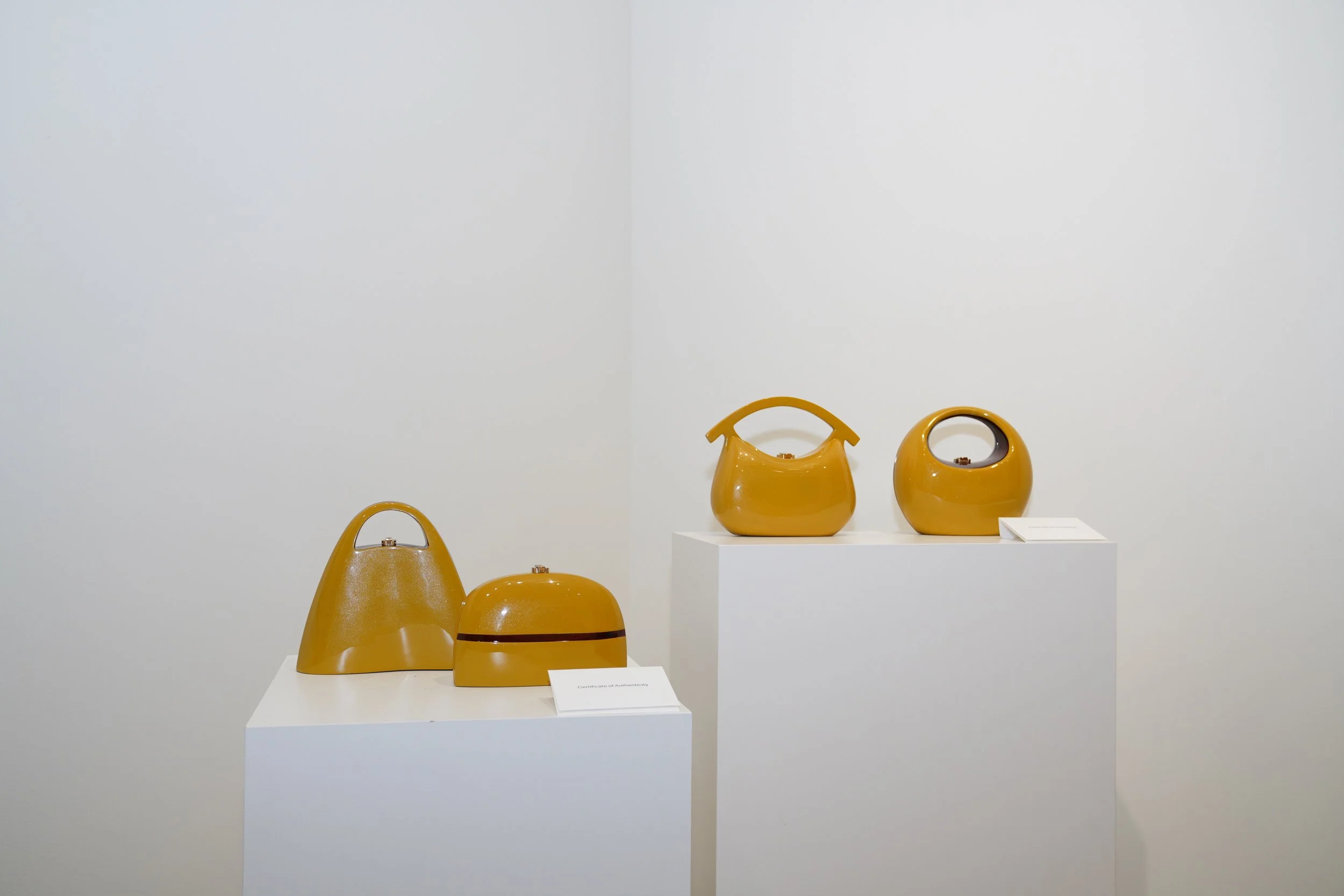 Art exhibit displaying four yellow-brown ceramic handbags and containers on two white pedestals against a plain white wall.