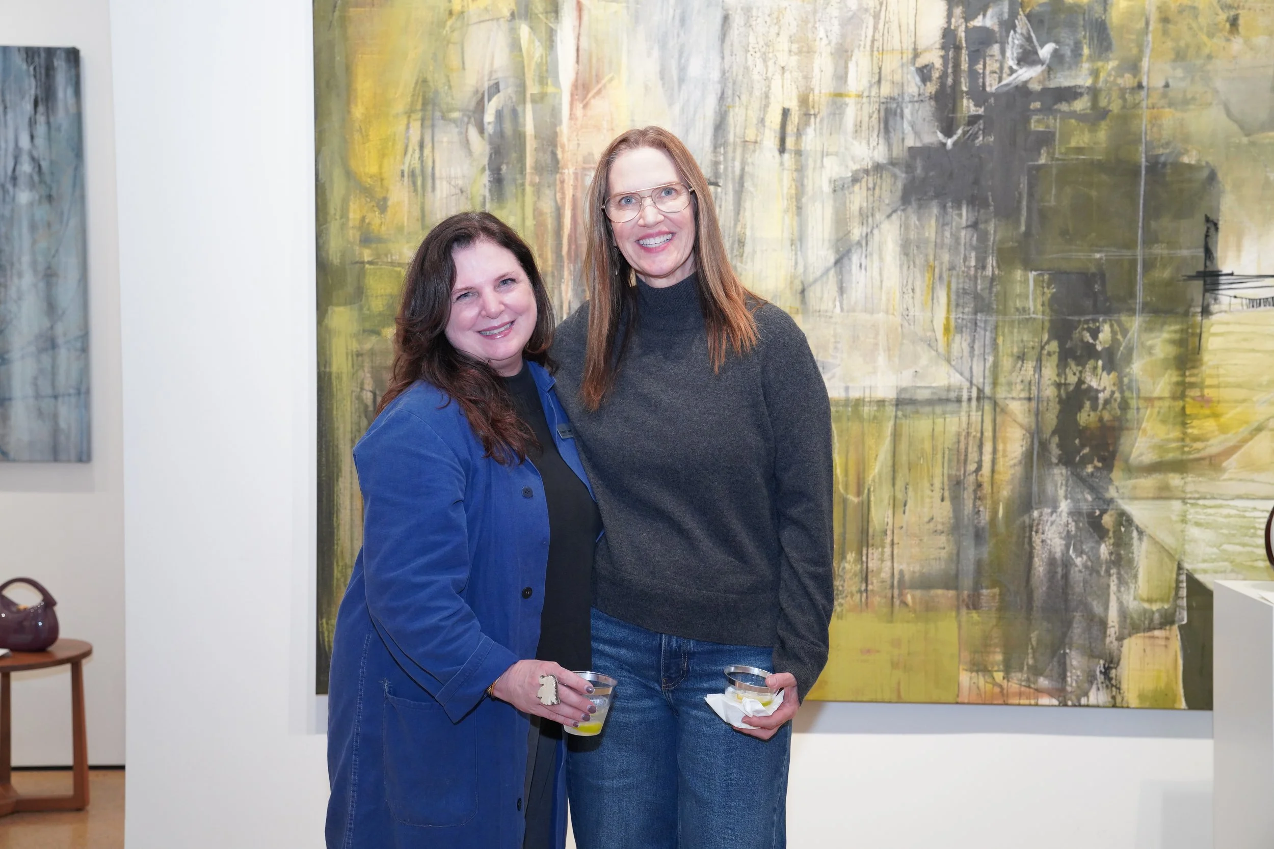 Two women standing together at an art gallery, smiling and holding drinks, with abstract paintings on the wall behind them.