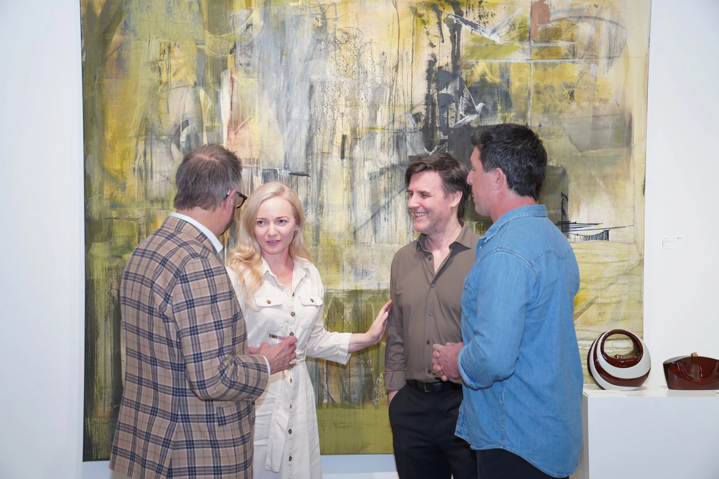 Four people engaged in conversation at an art gallery, with an abstract painting and pottery on a white pedestal in the background.