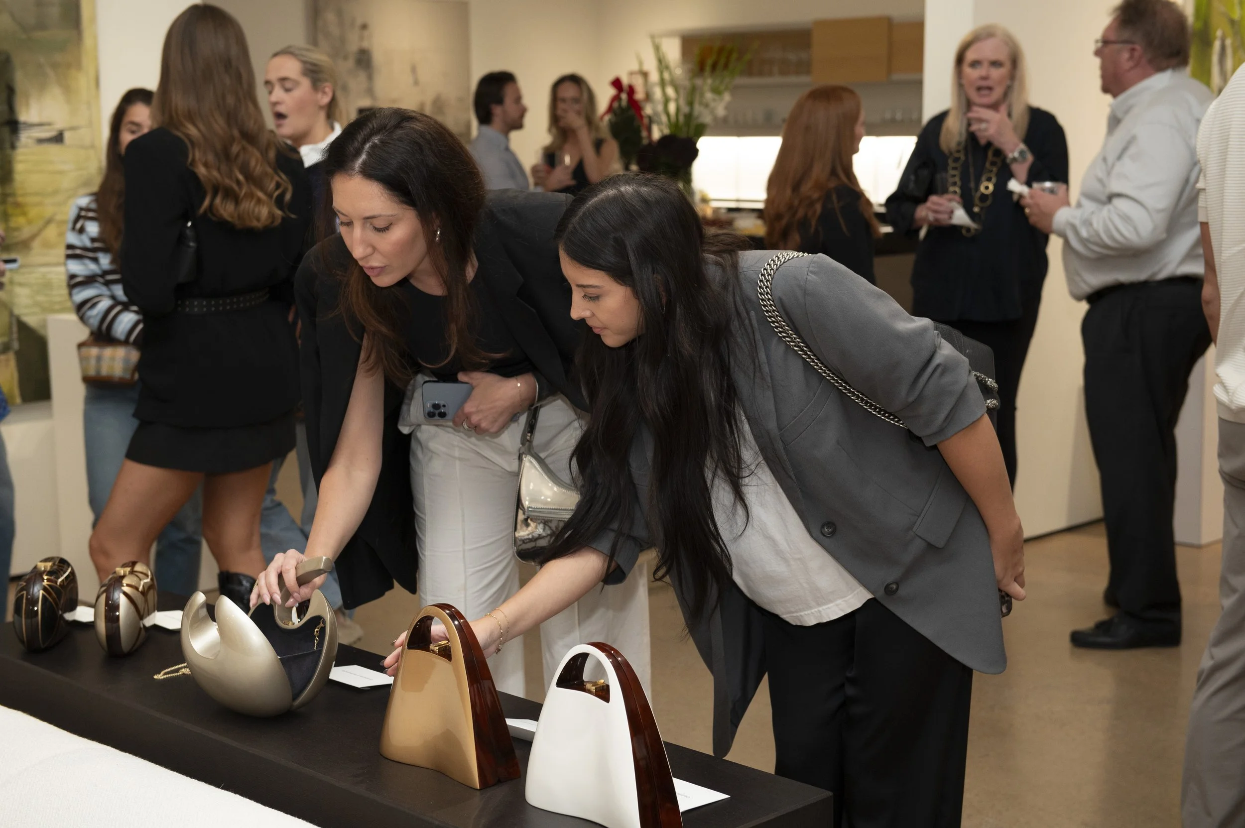 Two women are looking at designer handbags on display at an art gallery or boutique event, with other people in the background socializing.