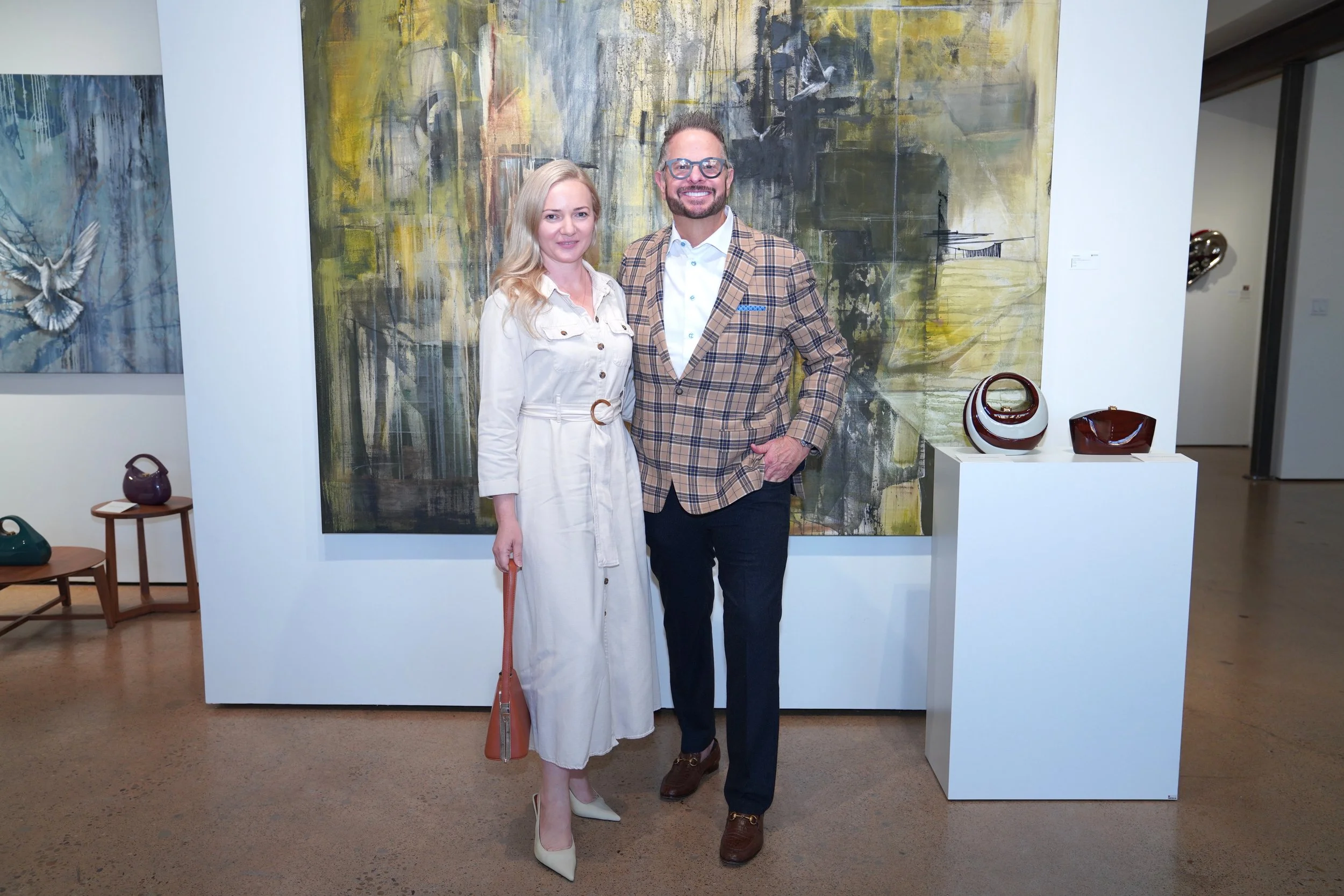 A man and woman standing together in an art gallery, smiling at the camera, with paintings and colorful vases displayed behind them.