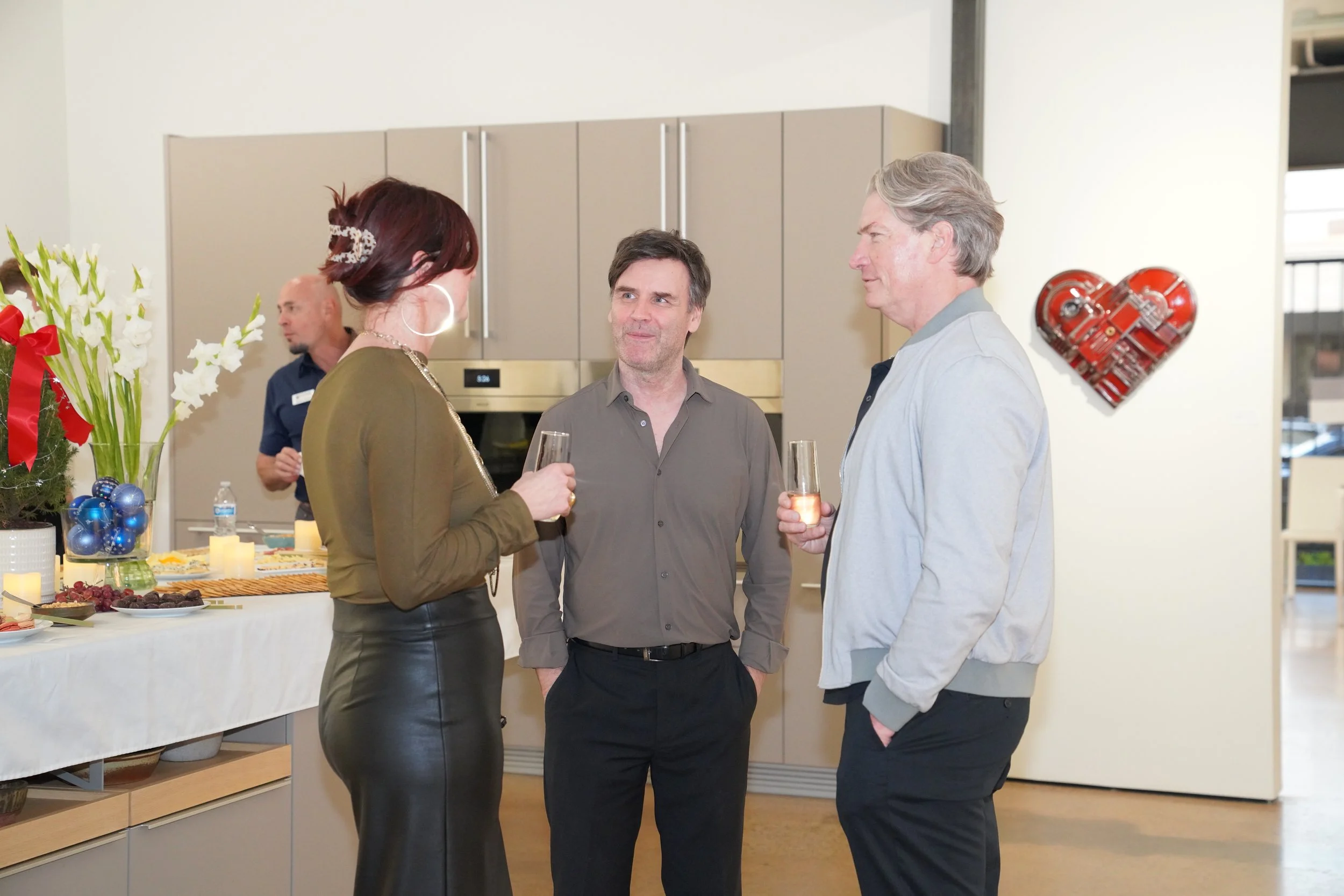 Three people talking and holding drinks at a social gathering. A woman with dark hair and hoop earrings is speaking to two men, one with gray hair and the other with dark hair, in a modern kitchen with a decorated table in the background.