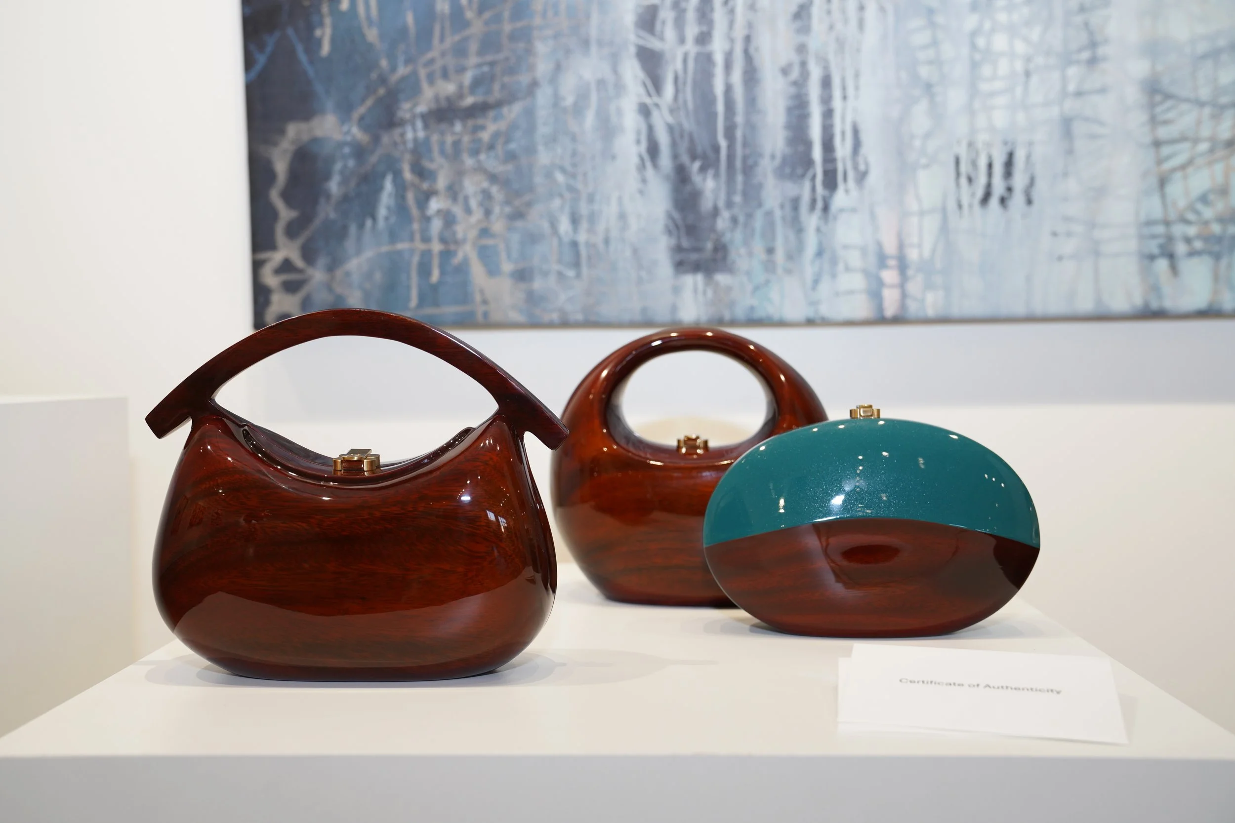 Three wooden handbags with curved handles displayed on a white pedestal in an art gallery, with abstract blue and white painting in the background.